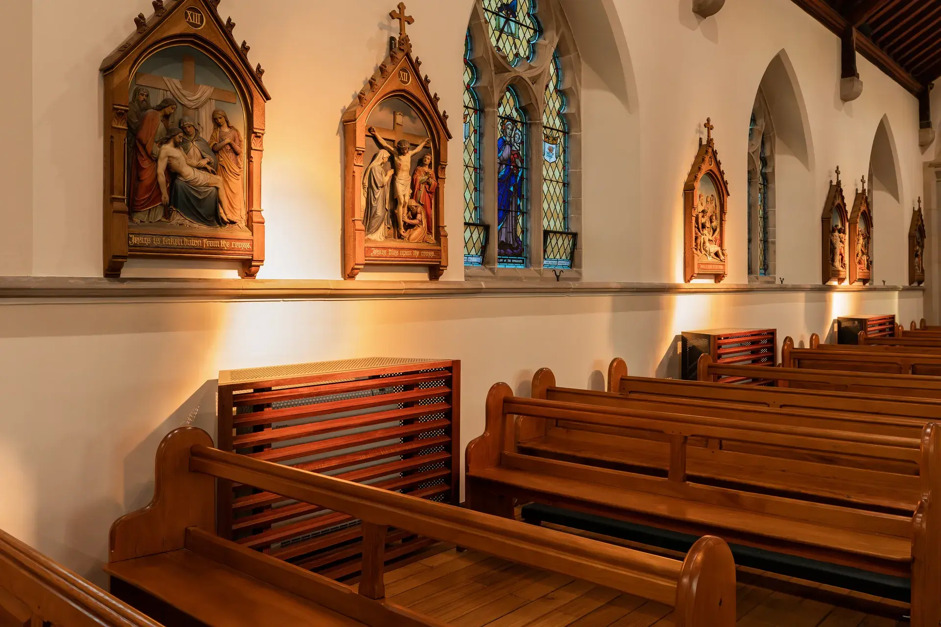 Interior of a cathedral featuring wooden pews, stained glass windows, and religious relief carvings mounted on the wall, all illuminated by warm, soft lighting.