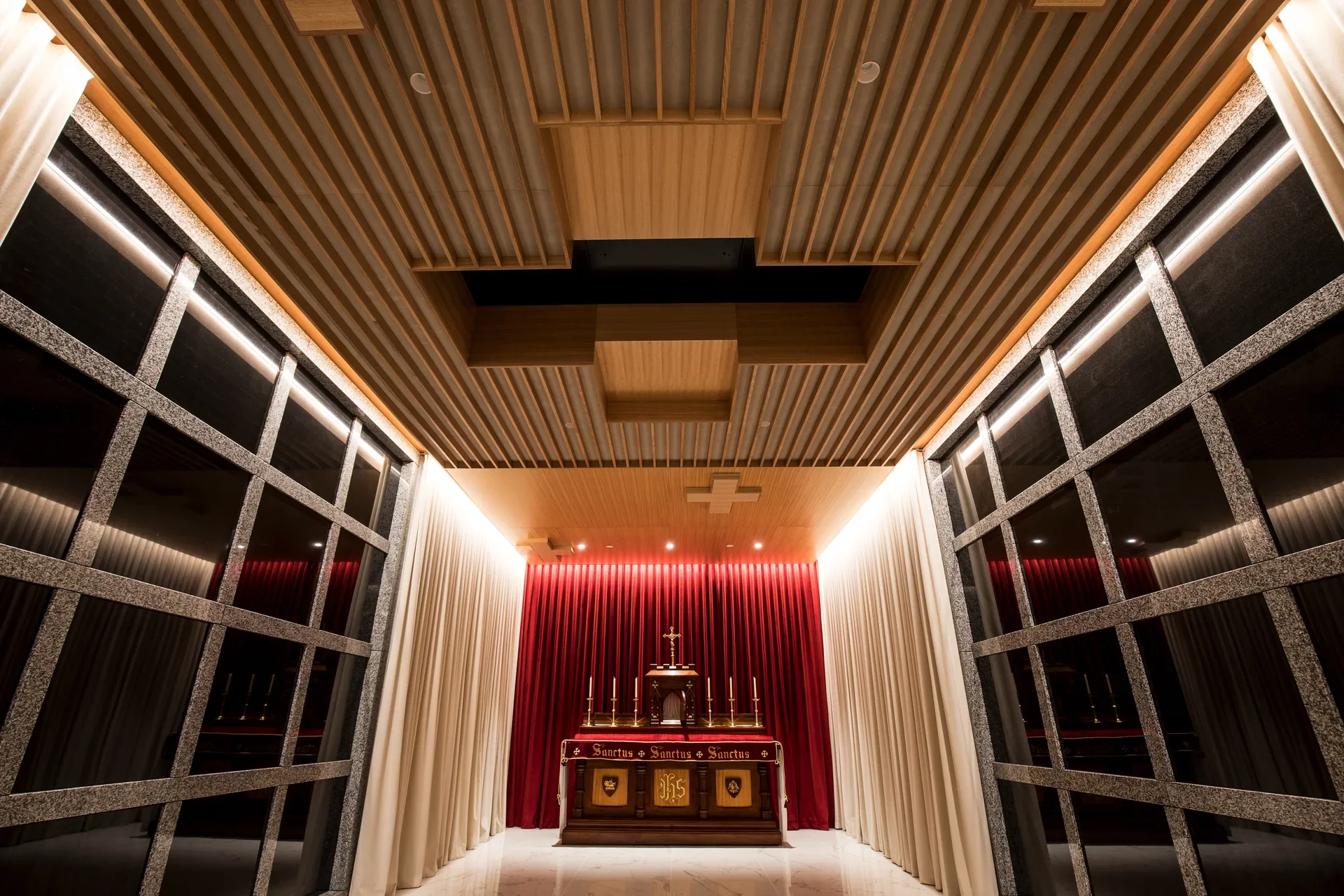 This small narthex features a highly detailed wooden slat ceiling with recessed sections and a cross motif. An ornate wooden altar stands against a vibrant red curtain, framed by long cream drapes and large grid-patterned glass walls on either side.