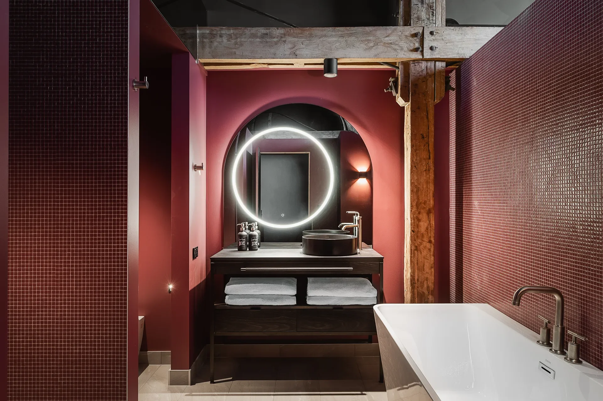 This bathroom features deep red walls and a large arched mirror with an integrated circular halo light. The vanity holds a black vessel sink and white towels, while a freestanding white bathtub is positioned in the foreground.