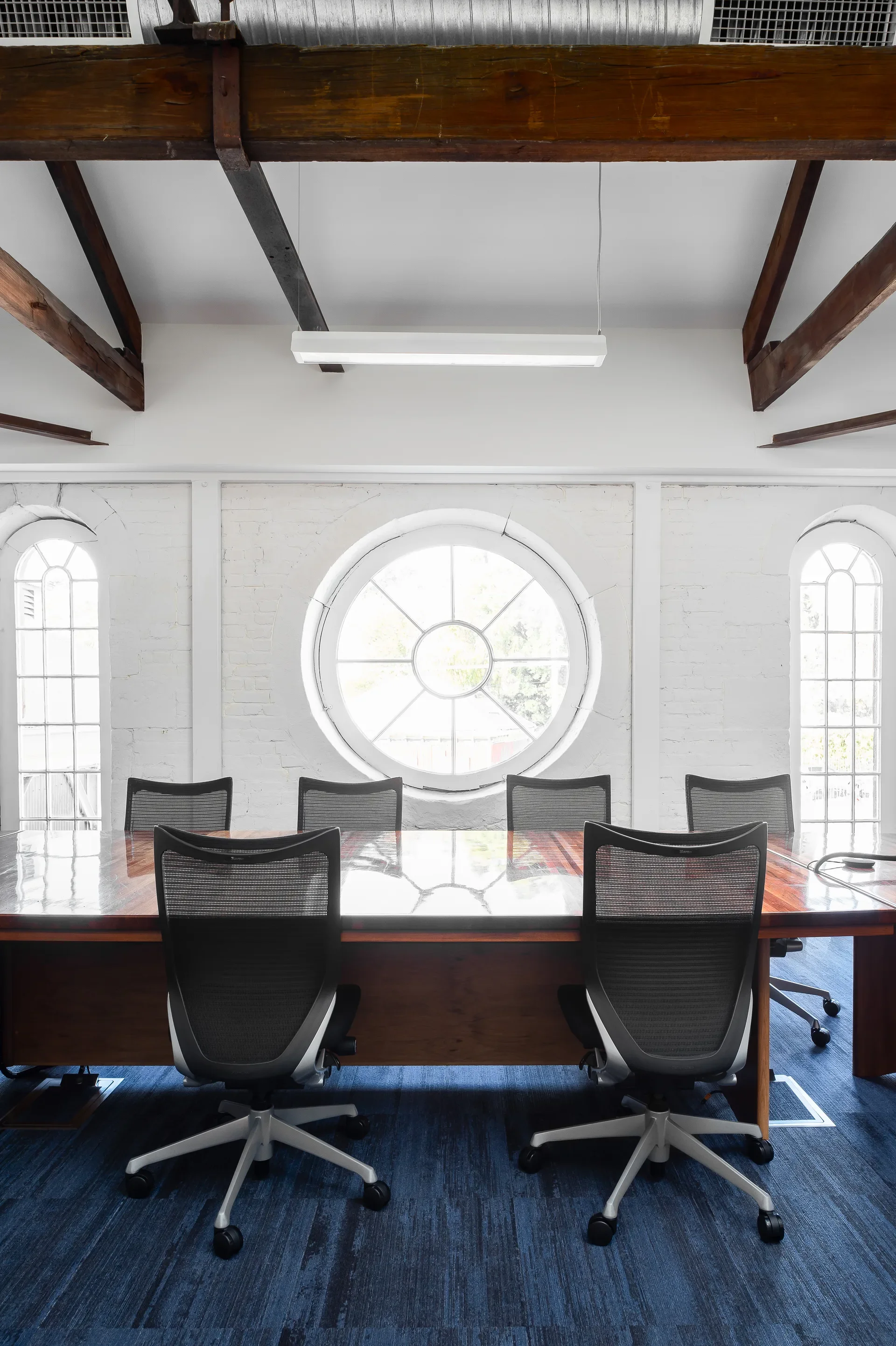 This conference room features a large circular window flanked by two narrow, arched windows set into a white brick wall. A polished wooden table is surrounded by modern mesh chairs, beneath a ceiling with exposed dark wood beams and a minimalist strip light.