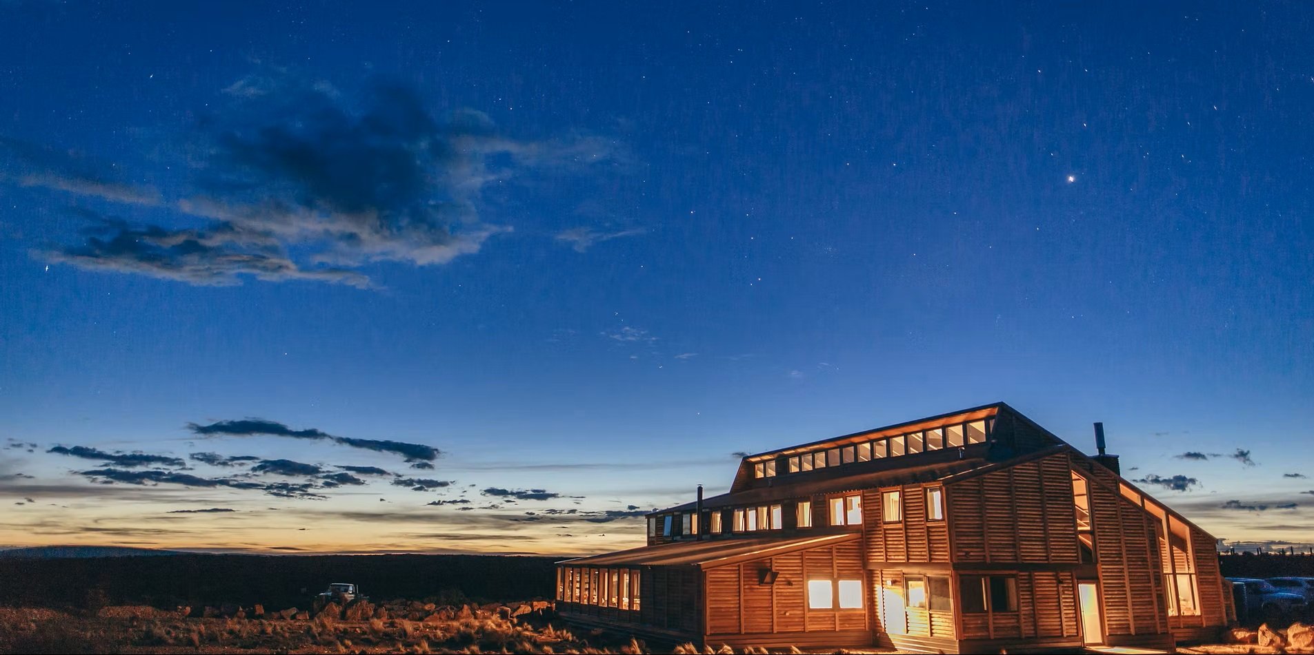 This large, multi-story timber lodge features a slanted roofline and glowing windows, set against a deep blue twilight sky filled with stars. The building is constructed with horizontal wood siding and includes a long row of clerestory windows along the top level.