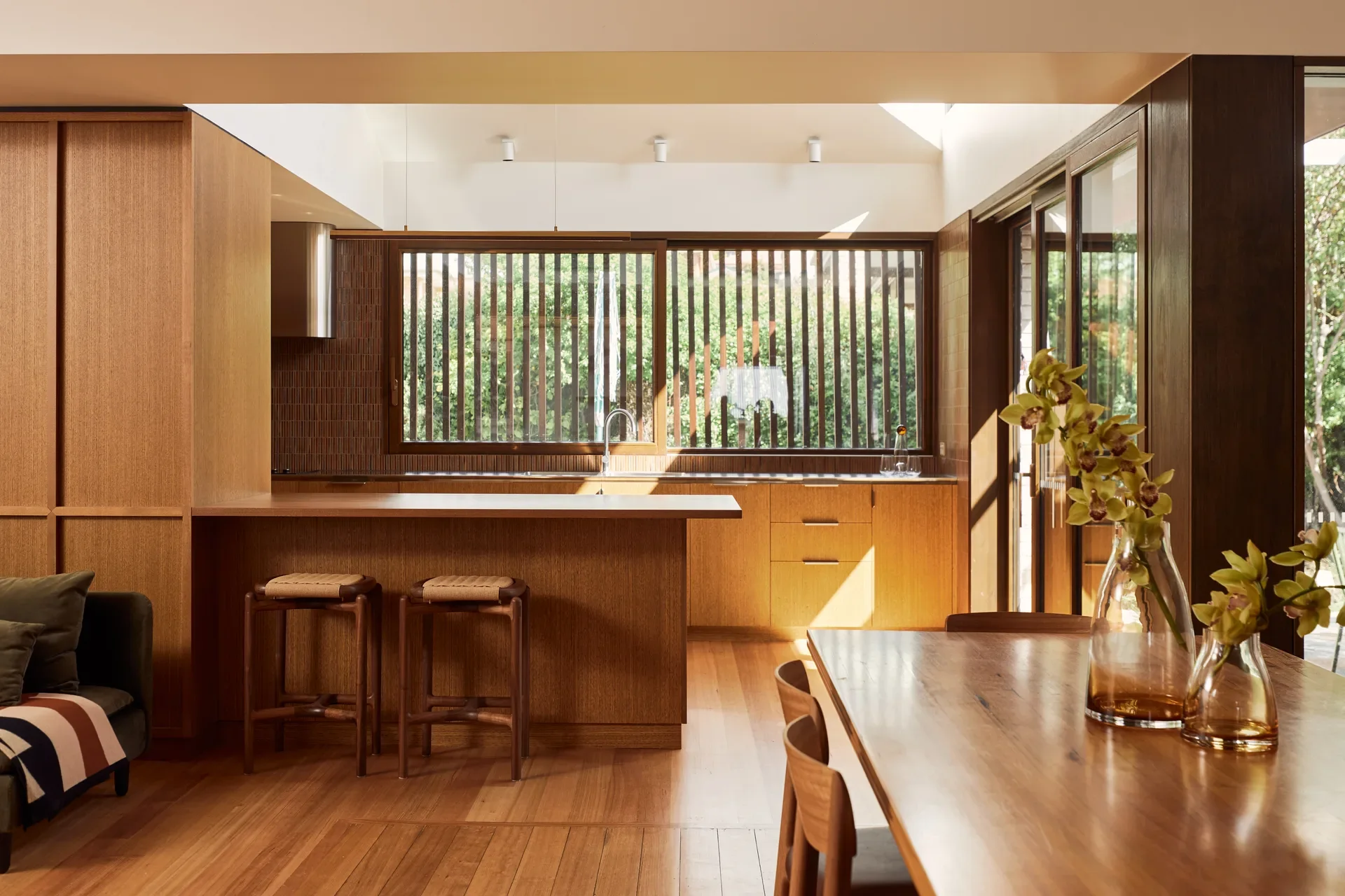 This warm, modern kitchen and dining area is dominated by rich wood tones and natural light. It features a minimalist wooden island and a large window with vertical timber slats that create a rhythmic pattern of light and shadow.