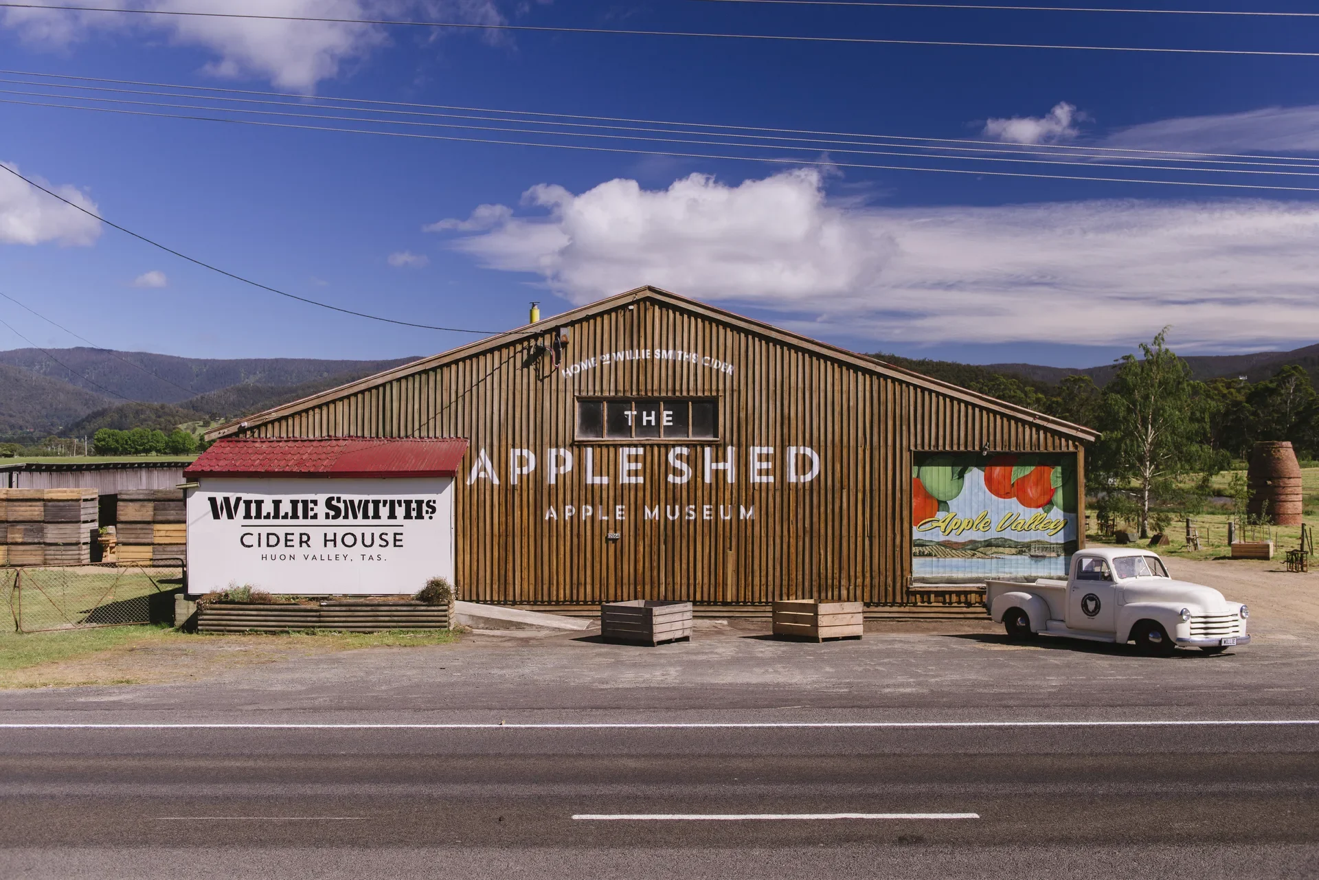 The Apple Shed and Apple Museum features vertical timber siding and large white signage. A vintage white pickup truck is parked in front of a colourful "Apple Valley" mural, next to the smaller "Willie Smith's Cider House" building.