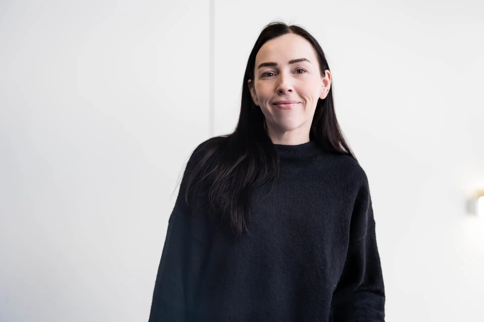 Portrait of a woman with long dark hair, smiling, wearing a black crewneck jumper, standing against a plain white wall.
