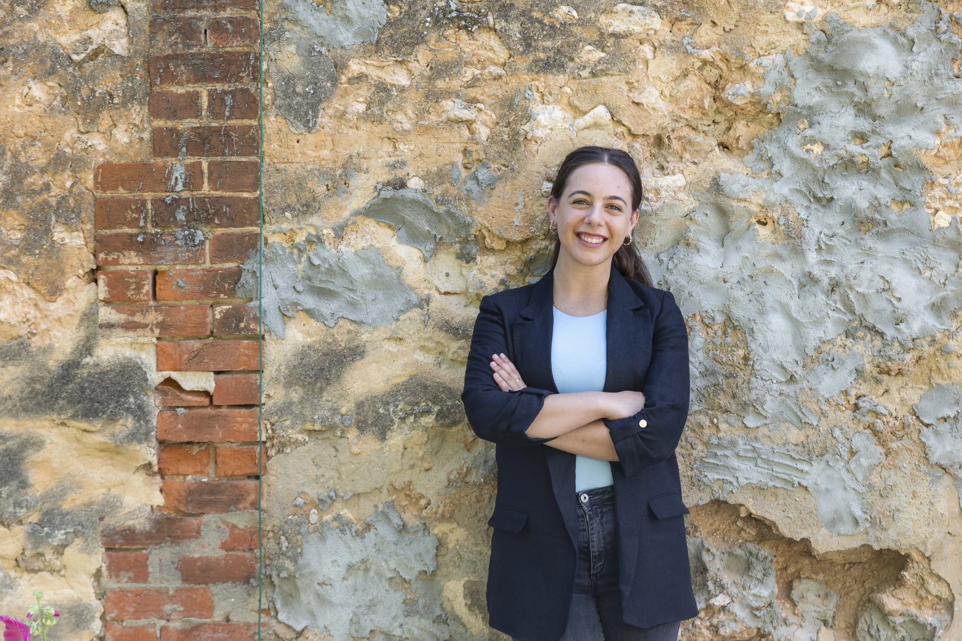 A professional headshot of Anastasia Mavrides standing against a textured, weathered wall combining exposed brick and rough plaster. She wears a black blazer over a light top, arms folded, smiling directly at the camera with a confident and approachable expression.