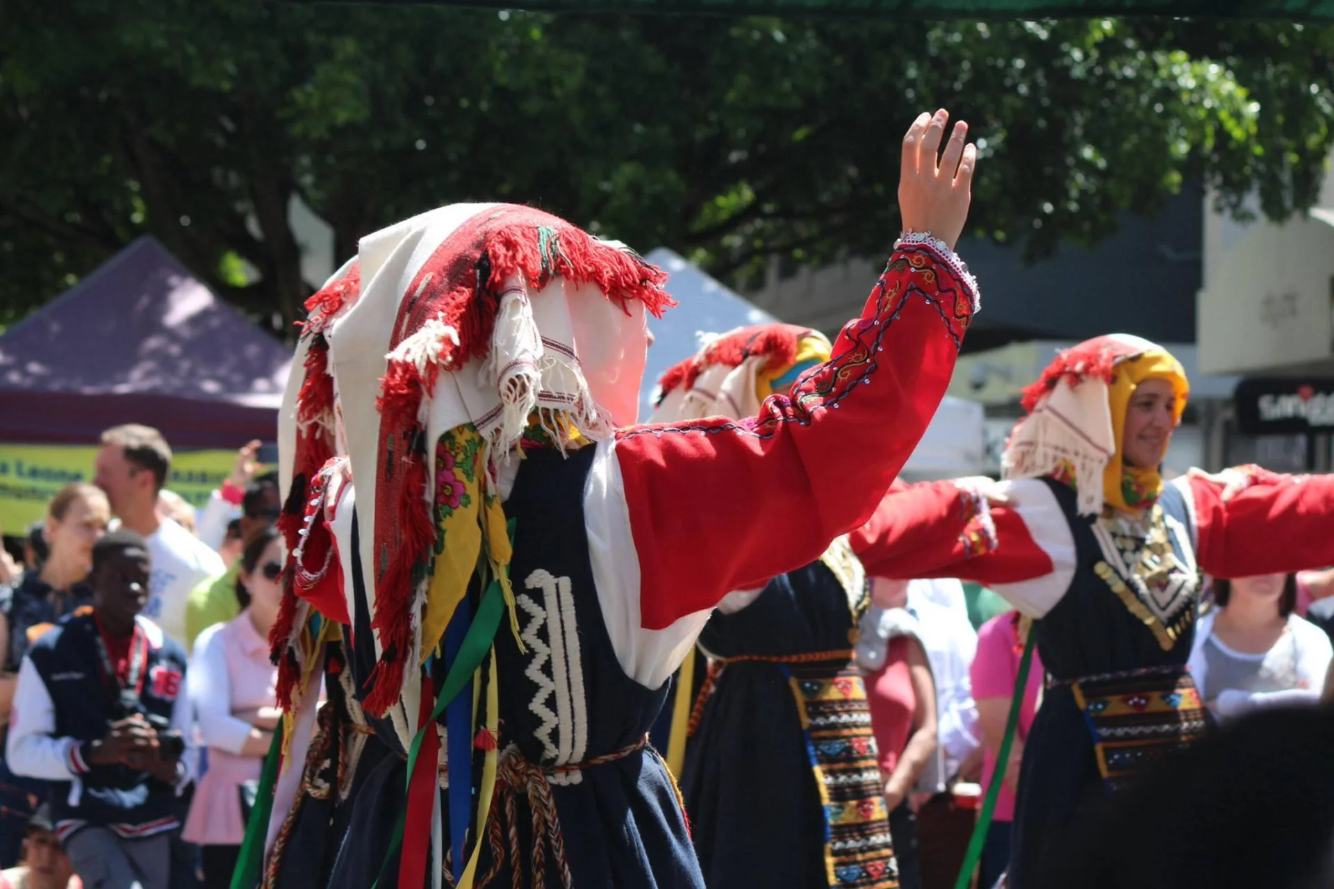 A group of people wearing traditional Greek garments perform a dance outdoors, dressed in richly coloured costumes with embroidered details and headscarves, surrounded by onlookers.
