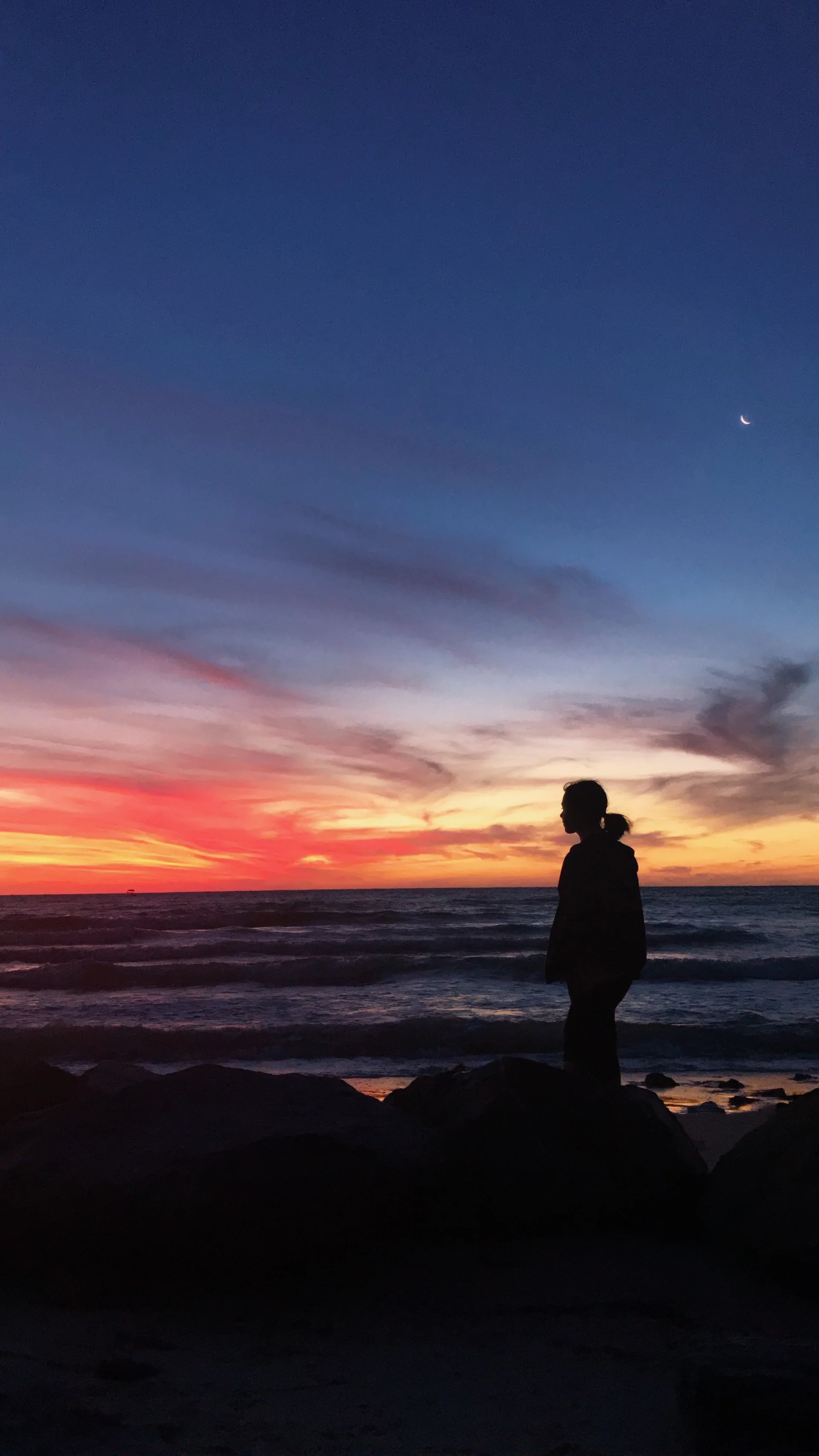 A silhouette of a person standing on rocks by the ocean at sunset, with a gradient sky of deep blue to warm orange and a crescent moon visible above.
