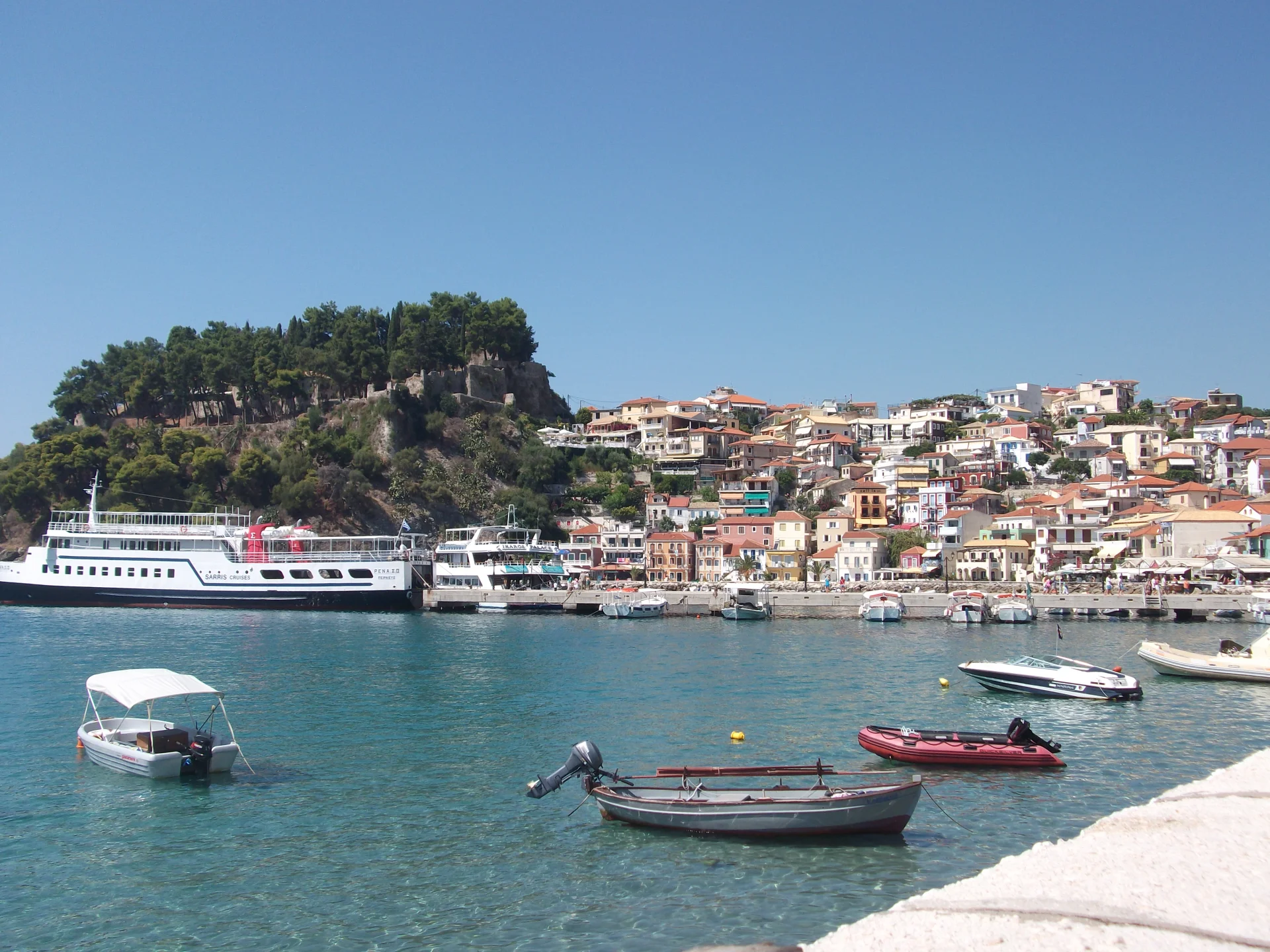 A coastal harbour scene in Greece with clear blue water, small boats in the foreground, and a hillside town of tightly packed buildings rising behind the waterfront.