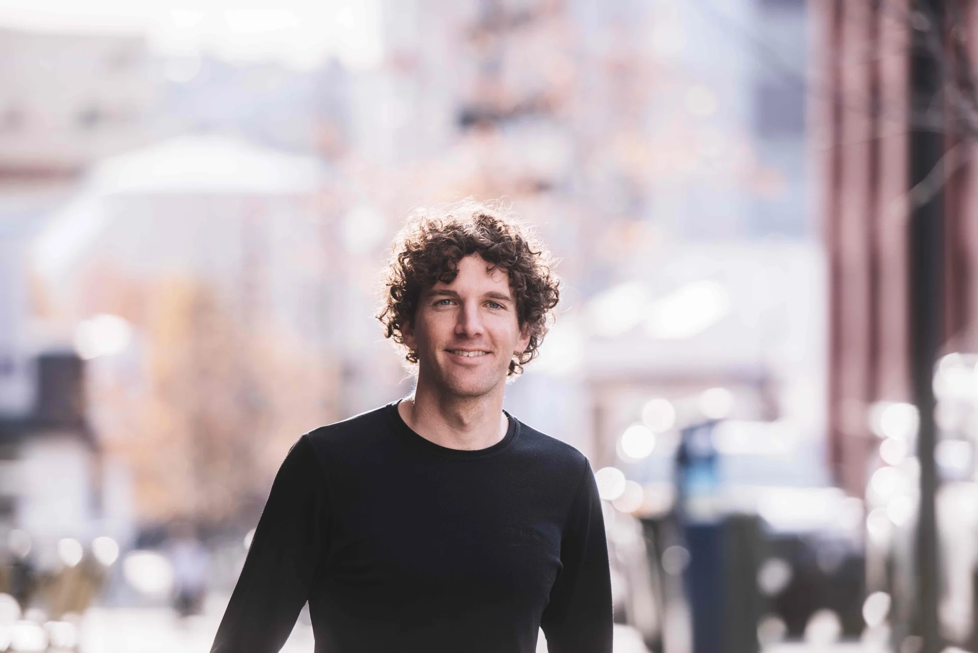 A professional headshot of Andrew Geeves standing outdoors on a softly blurred city street. He wears a black long-sleeve top, with short curly brown hair, smiling directly at the camera with a relaxed and approachable expression.