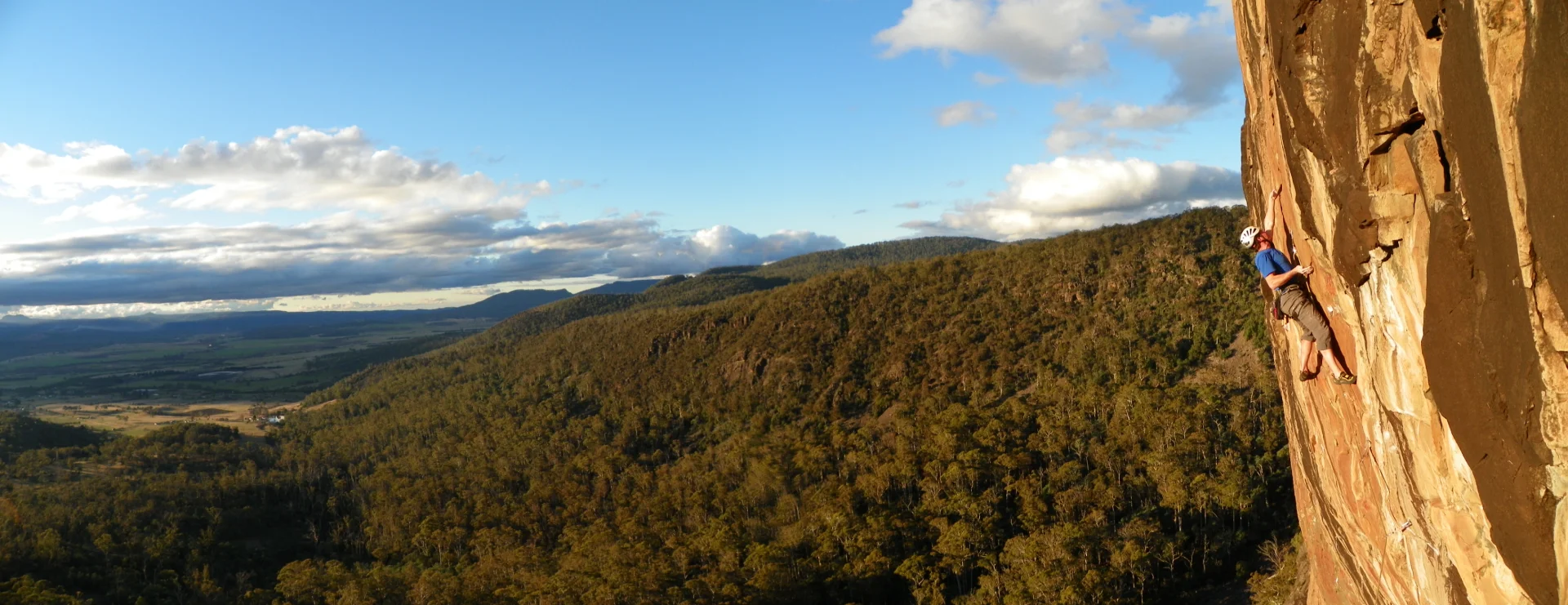 A panoramic image of Andrew rock climbing a steep cliff face above a vast forested landscape, with distant hills and a wide sky in the background.