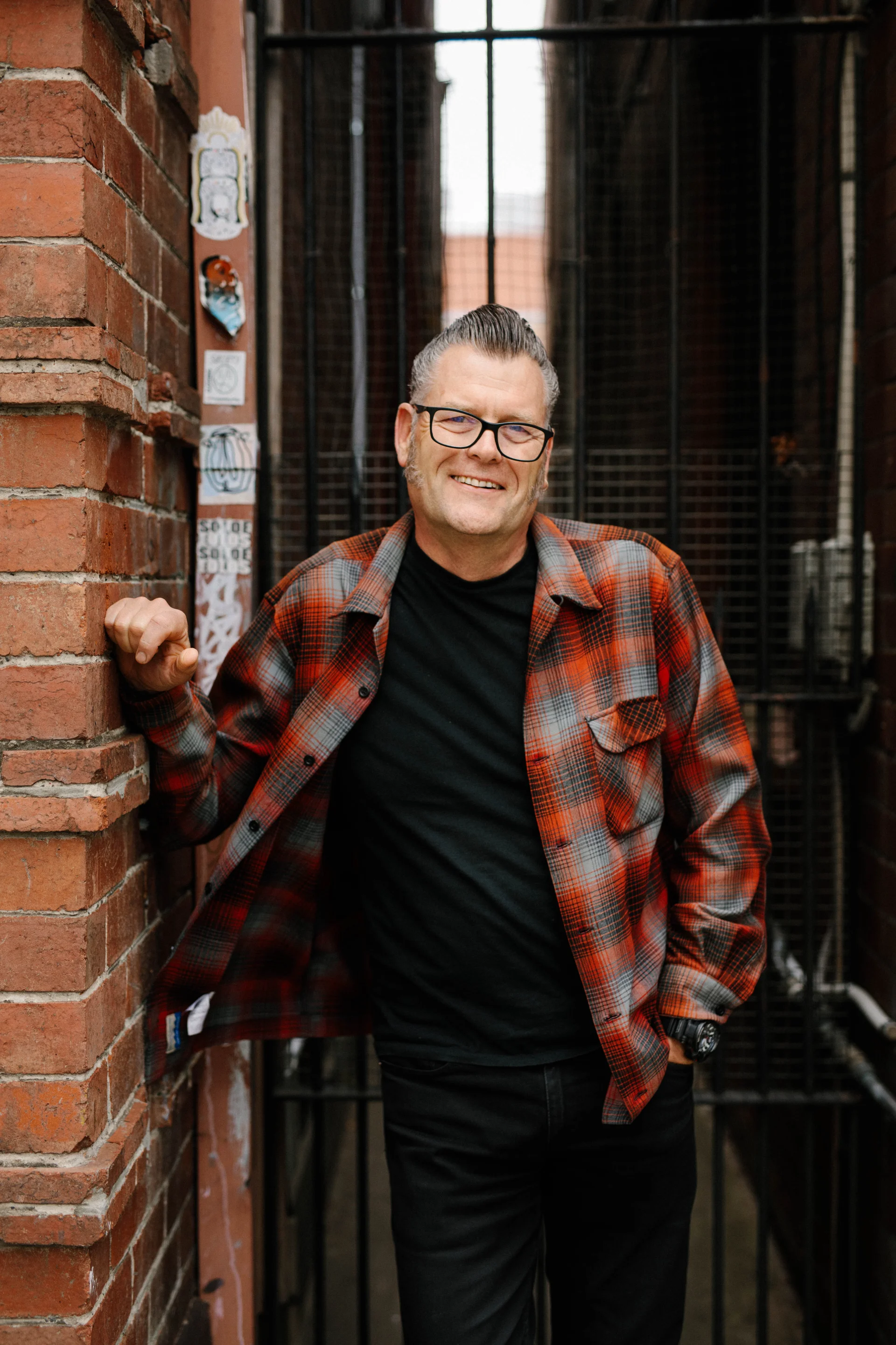 A professional headshot of Andrew Grimsdale standing outdoors beside a brick wall and urban backdrop. He wears a red and grey plaid overshirt over a black top, with glasses and neatly styled hair, smiling warmly at the camera with a relaxed and approachable expression.