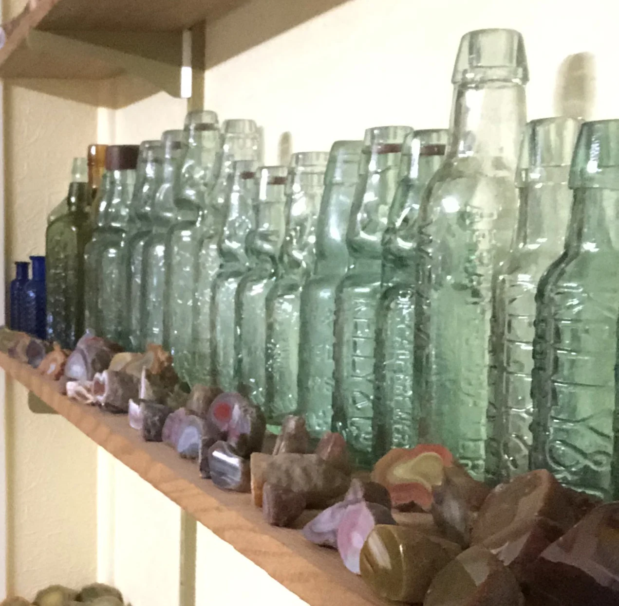 A row of translucent glass bottles in soft green and blue tones displayed on a wooden shelf, accompanied by an assortment of small stones and objects.