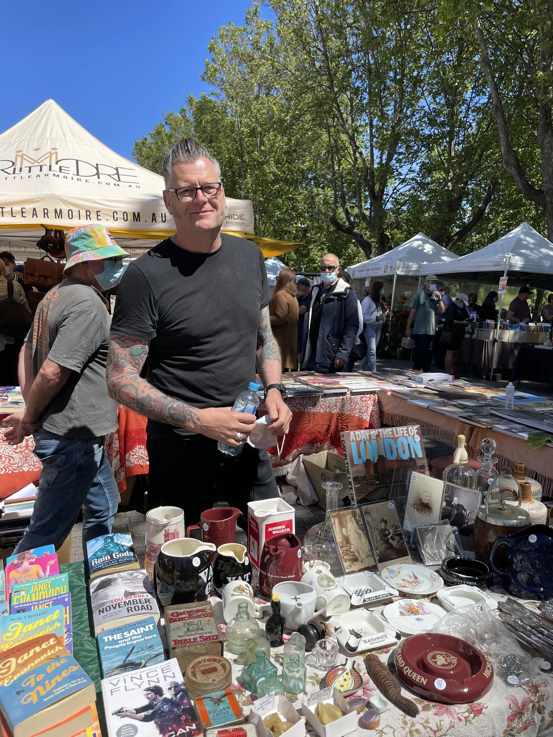 Andrew standing at a Salamanca Market stall surrounded by vintage items, books, and ceramics, engaging with objects in a lively and informal setting. Andrew is wearing a short sleeved grey shirt, with colourful tattoos on his arms visible.