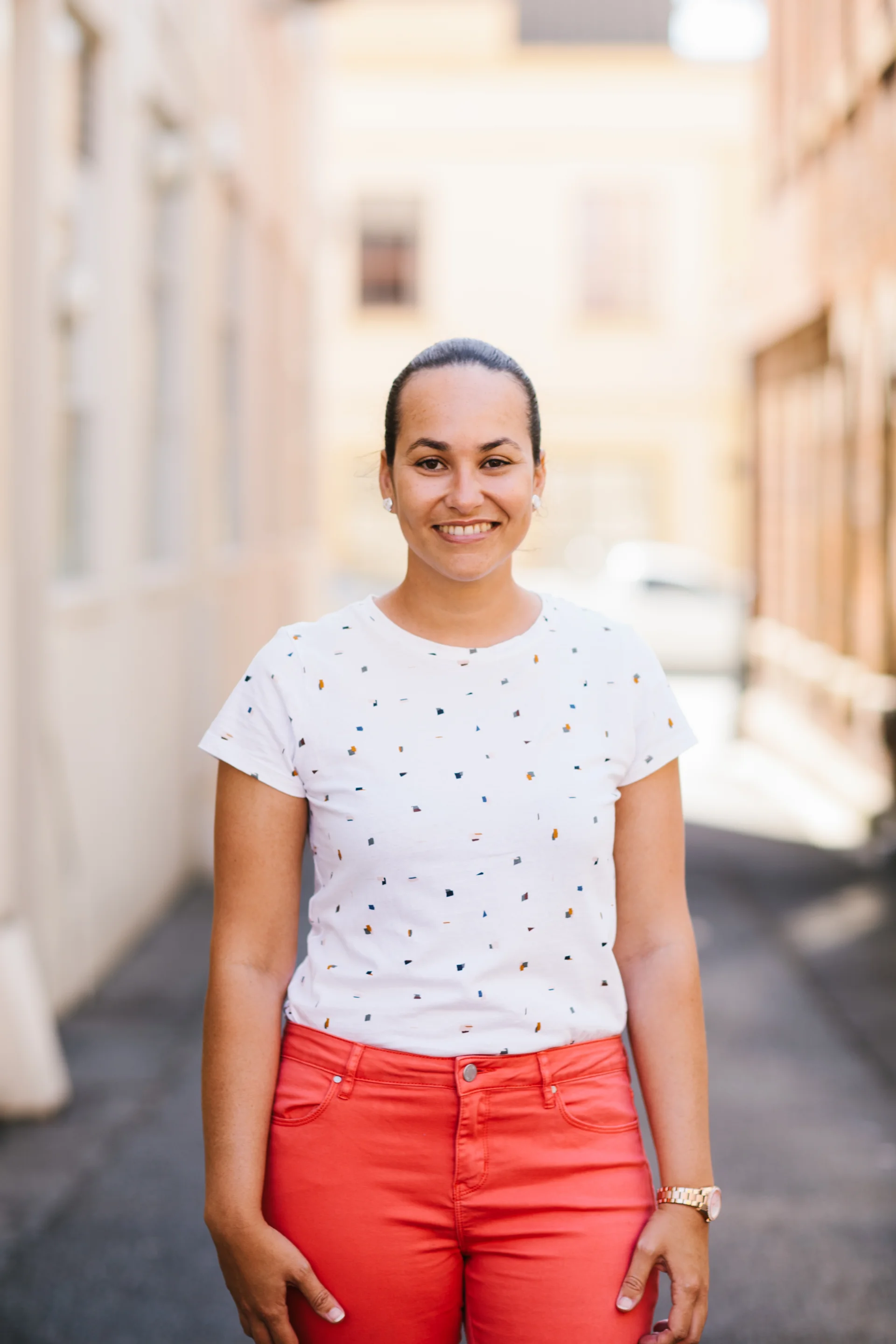 A professional headshot of Anna Alvis standing outdoors in a softly lit urban setting. She wears a light, patterned short-sleeve top, with her hair neatly tied back, smiling directly at the camera with a warm and approachable expression.