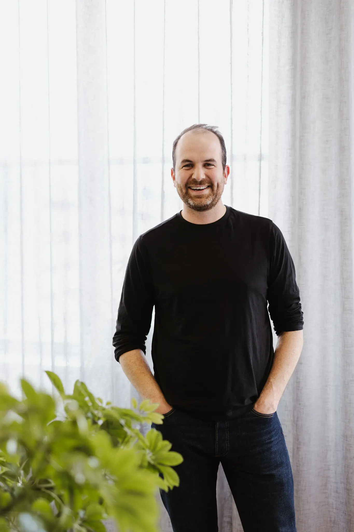 A professional headshot of Ben Sheehan standing indoors against a softly lit, sheer curtain backdrop. He wears a black long-sleeve top, with short hair and beard, smiling directly at the camera with a relaxed and friendly expression.
