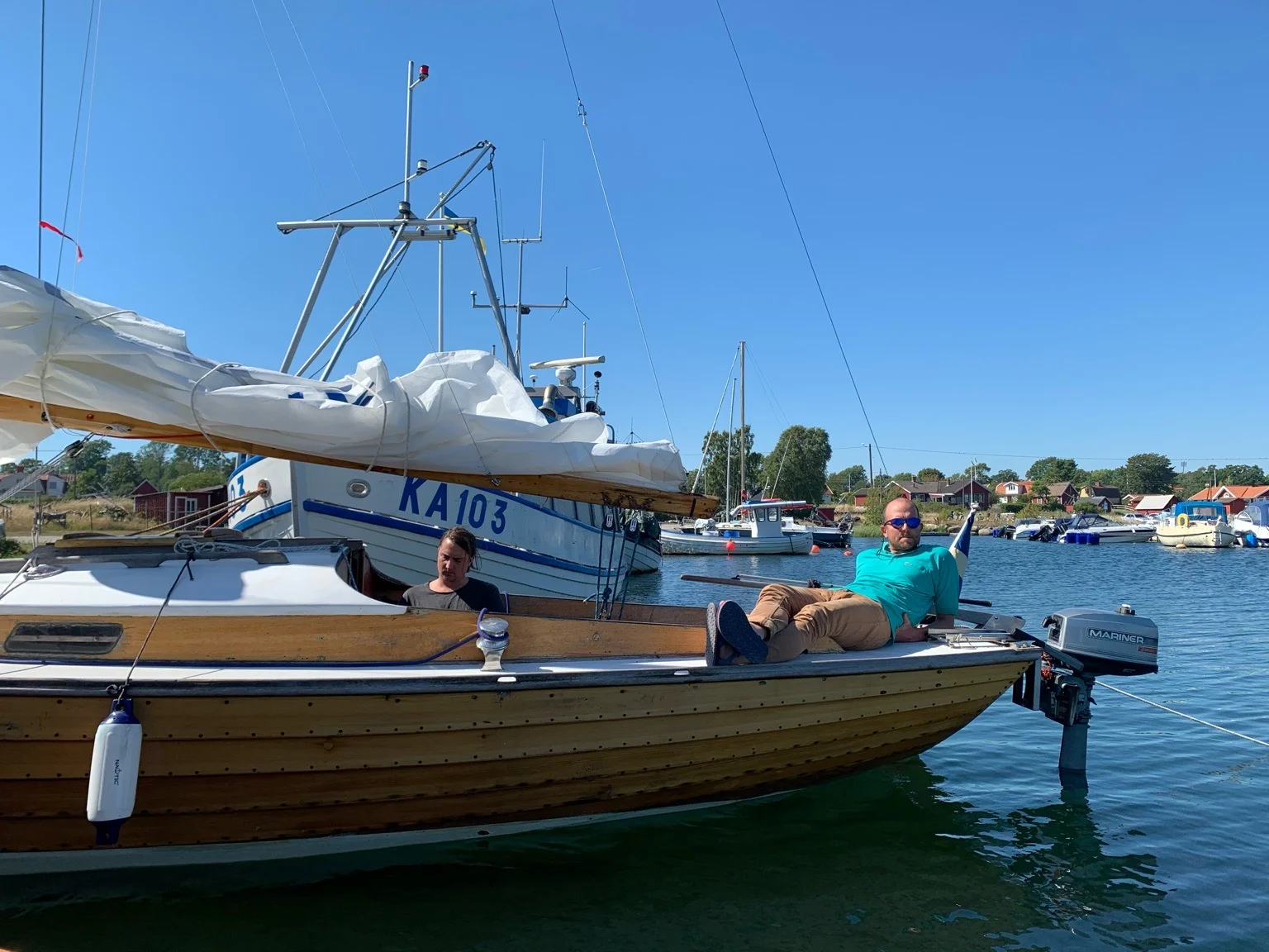 Ben laying casually on a small wooden boat in a harbour setting, surrounded by other moored vessels under clear blue skies.