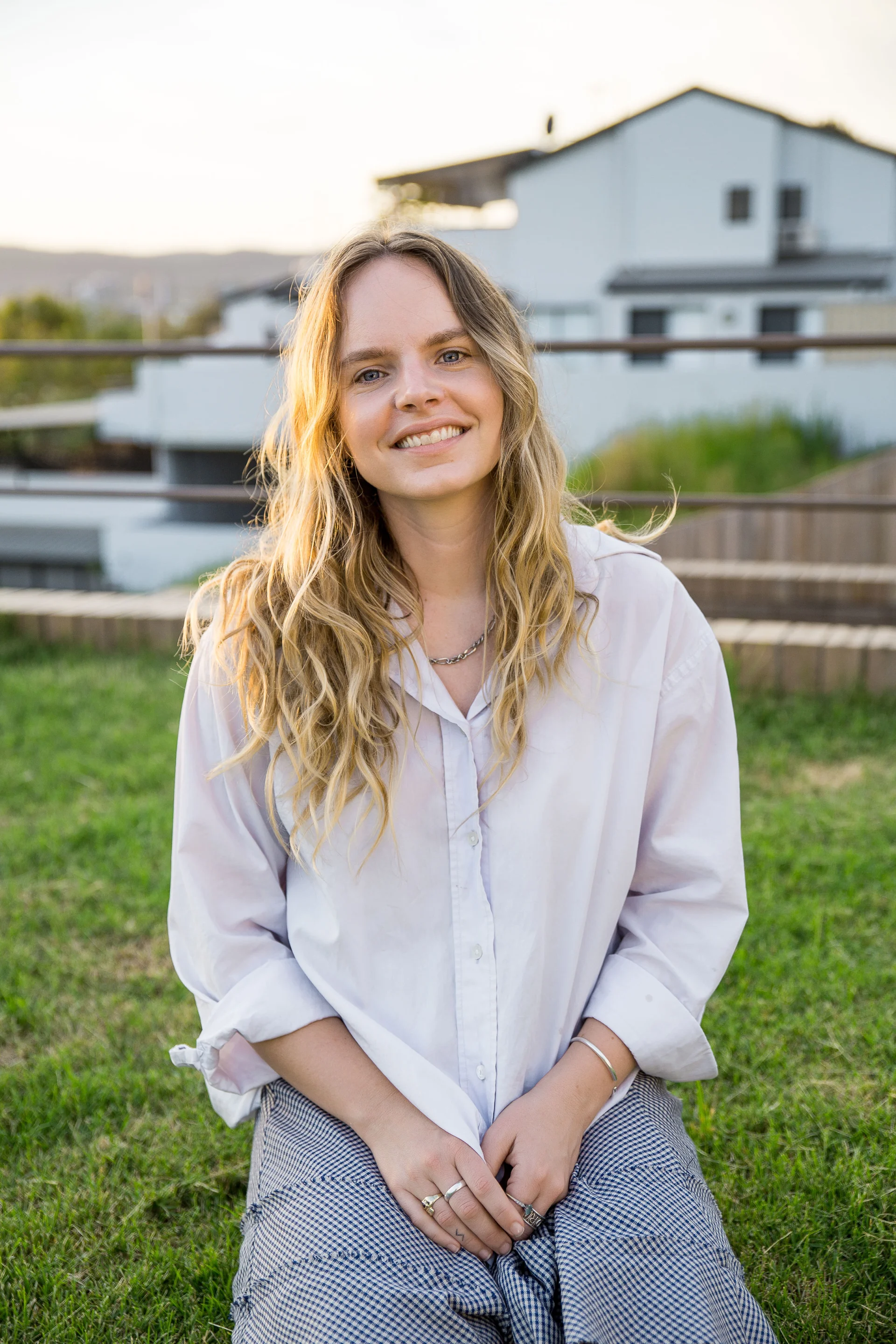 A portrait of Claudia Reinhardt smiling warmly outdoors, with long wavy blonde hair and a light shirt, softly lit by golden-hour sunlight against a blurred residential background.