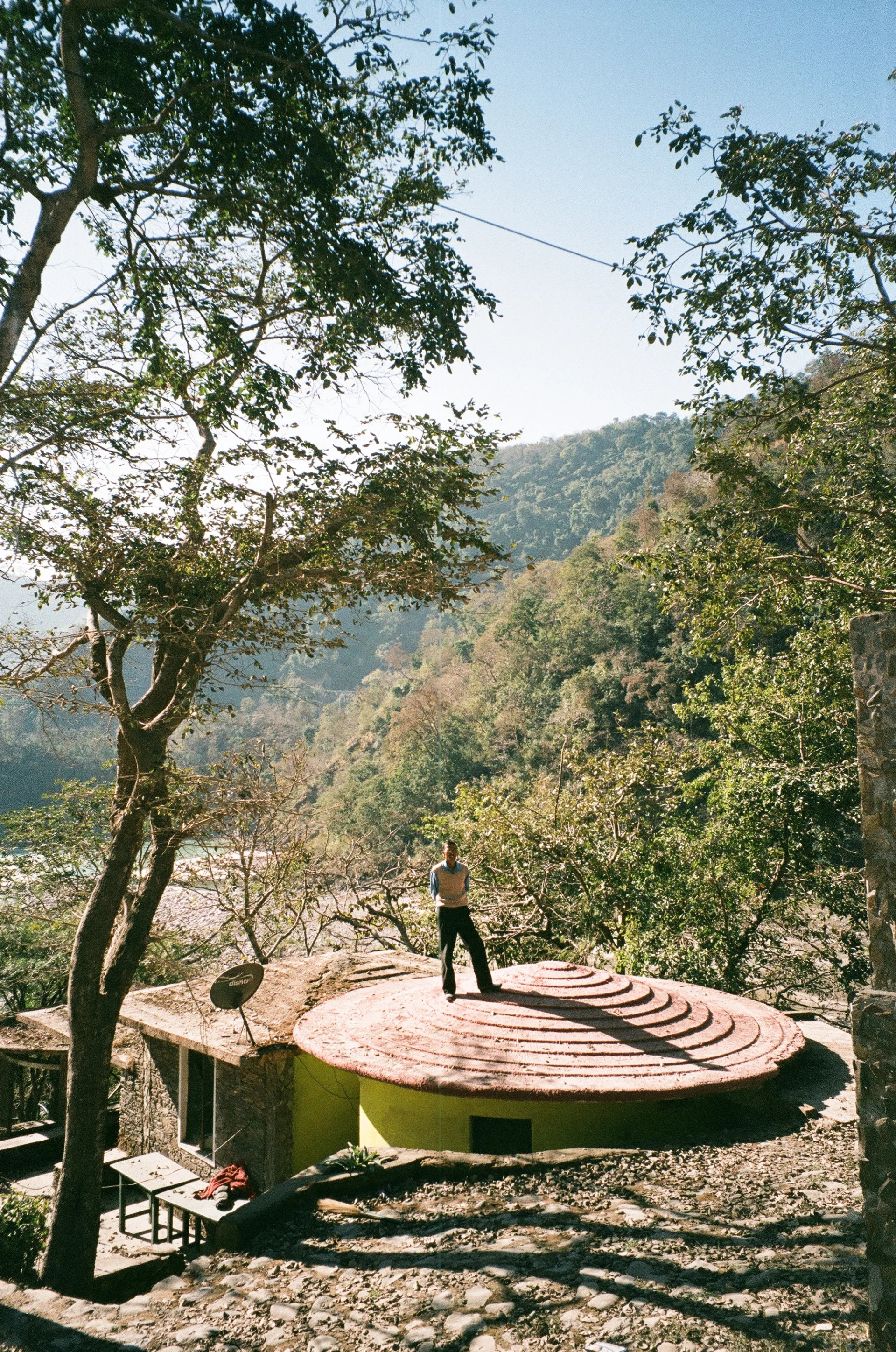 A scenic landscape of a forested hillside with a circular rooftop structure in the foreground, where a person stands on top, surrounded by trees and layered terrain.