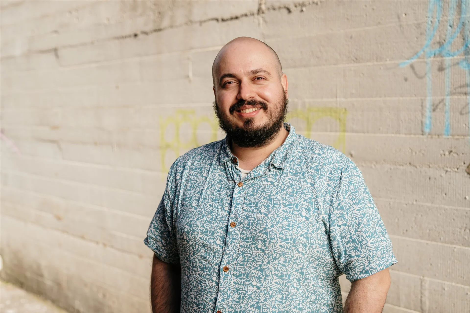 A portrait of Dmitry Troyanovsky smiling, with a shaved head and dark beard, wearing a patterned short-sleeve blue shirt, standing against a concrete wall with faint graffiti.