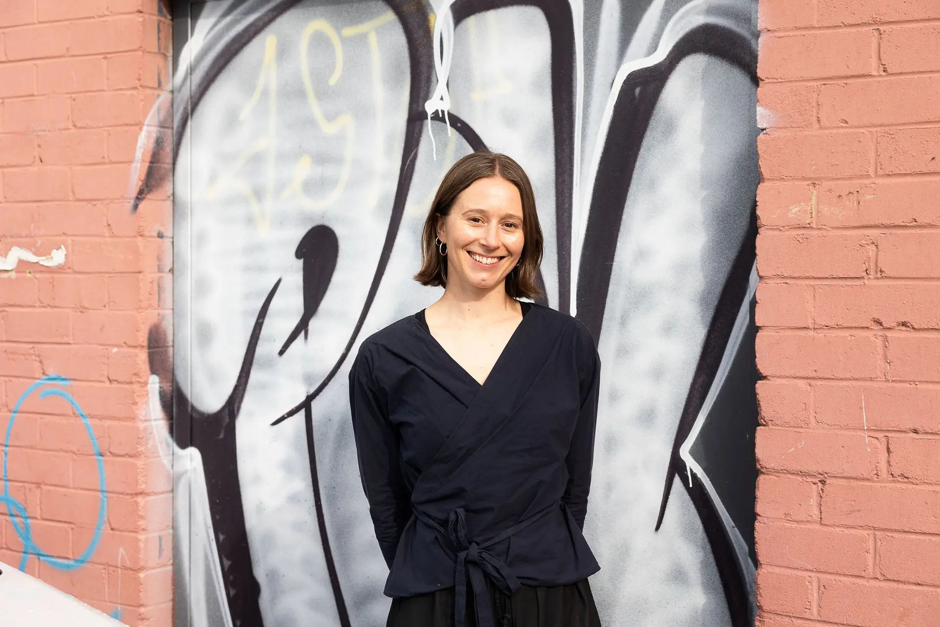 A photo of Dominique Petterwood standing against a brick background with graffiti behind her. She is wearing a black dress and is smiling at the camera.