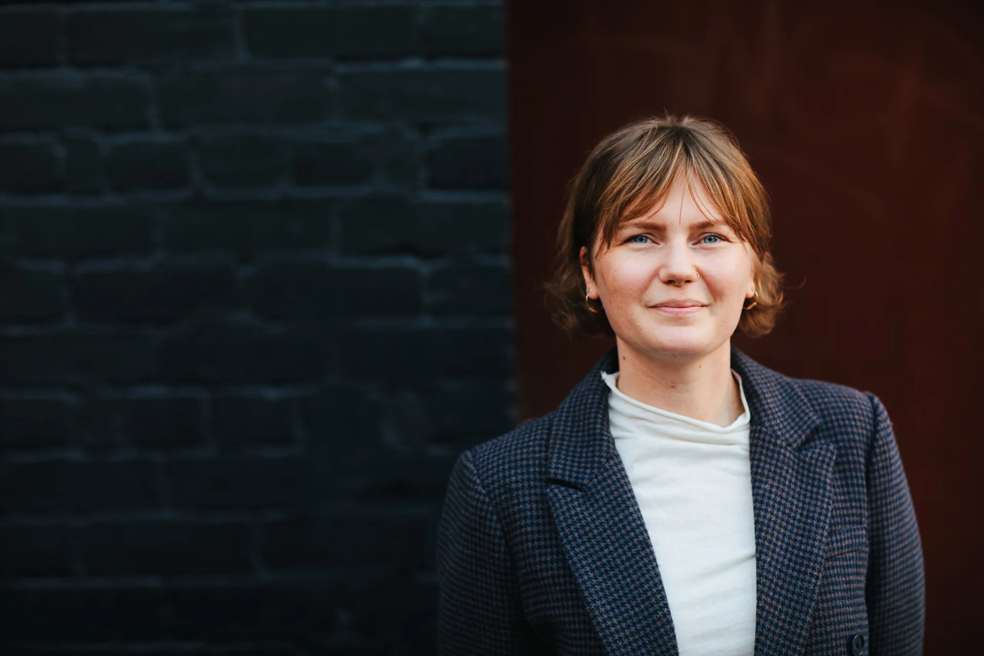 A professional headshot of Ebony Hurd standing outdoors against a dark red and black wall. She wears a dark blue blazer over a light top, with neat short hair, looking directly at the camera with a calm and composed expression.