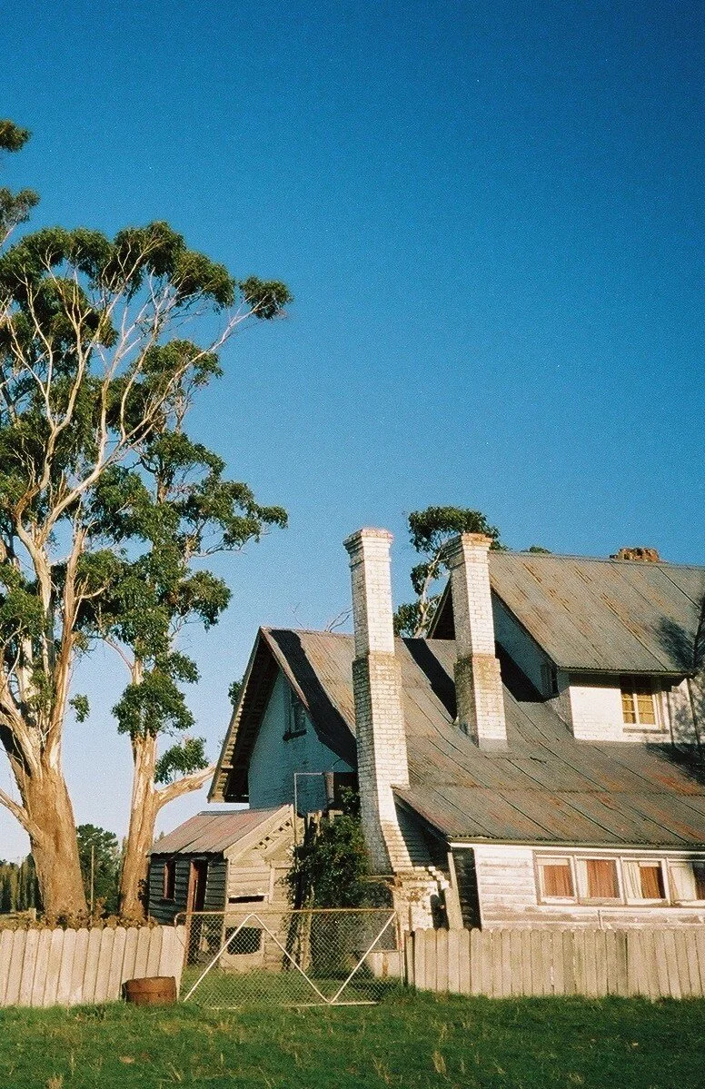 A beautiful weathered house with a corrugated roof and two tall chimneys, set behind a fence with trees beside it under a clear blue sky.