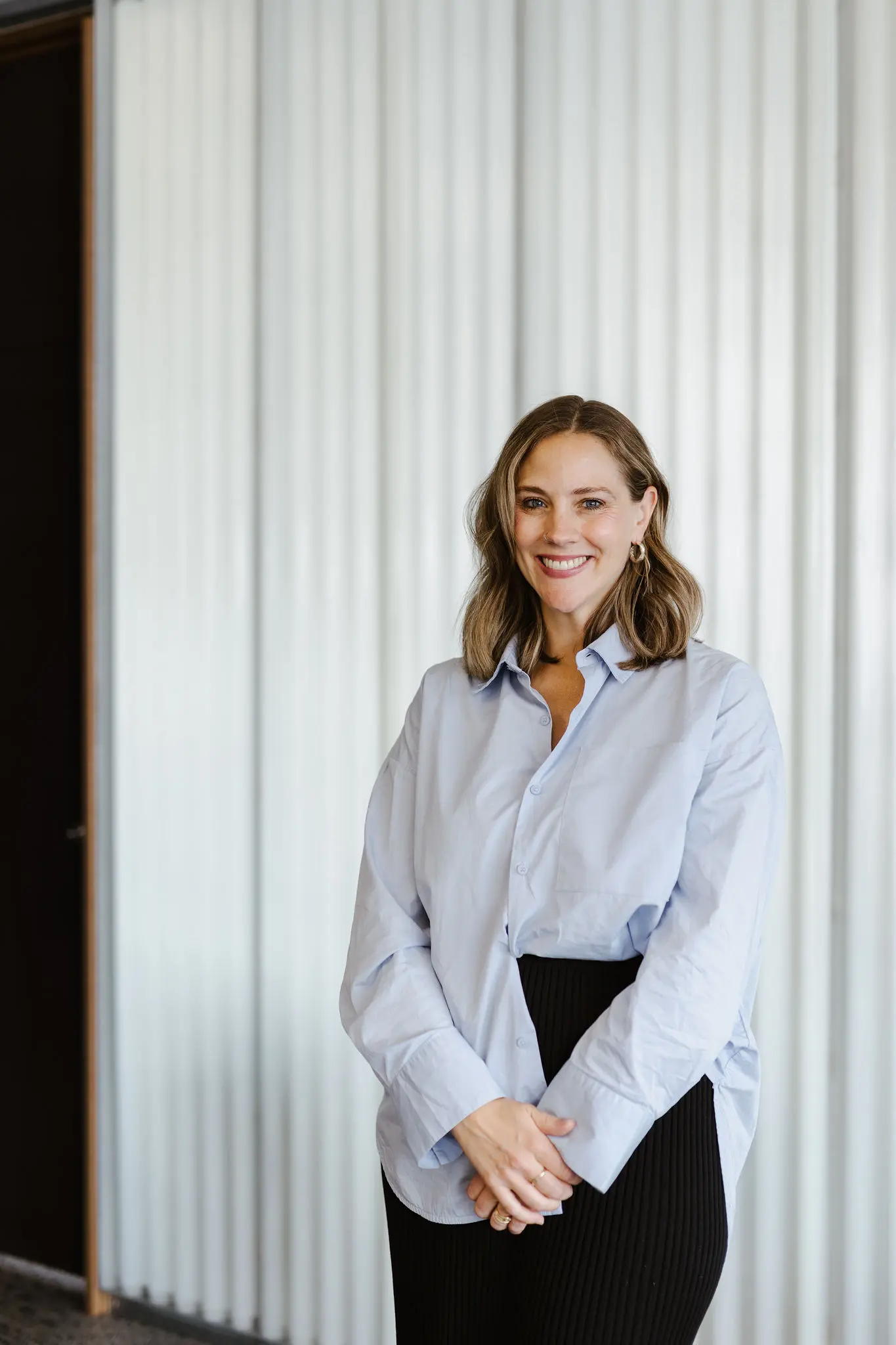A professional headshot of Georgina Russell standing indoors in the Hobart studio against a polycarbonate backdrop. She wears a light blue button-up shirt with her hair styled in loose waves, smiling directly at the camera with a friendly and confident expression.
