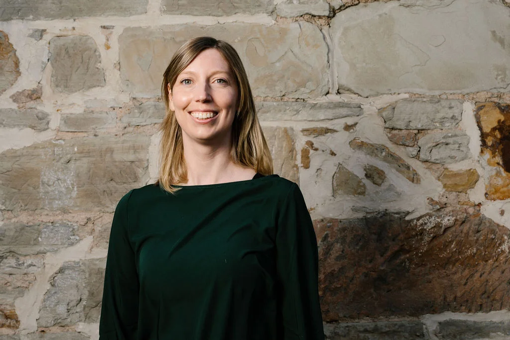 A professional headshot of Imogen Siberry standing in front of a stone wall. She wears a dark green long-sleeve top, with straight shoulder-length blonde hair, smiling directly at the camera with a friendly and approachable expression.