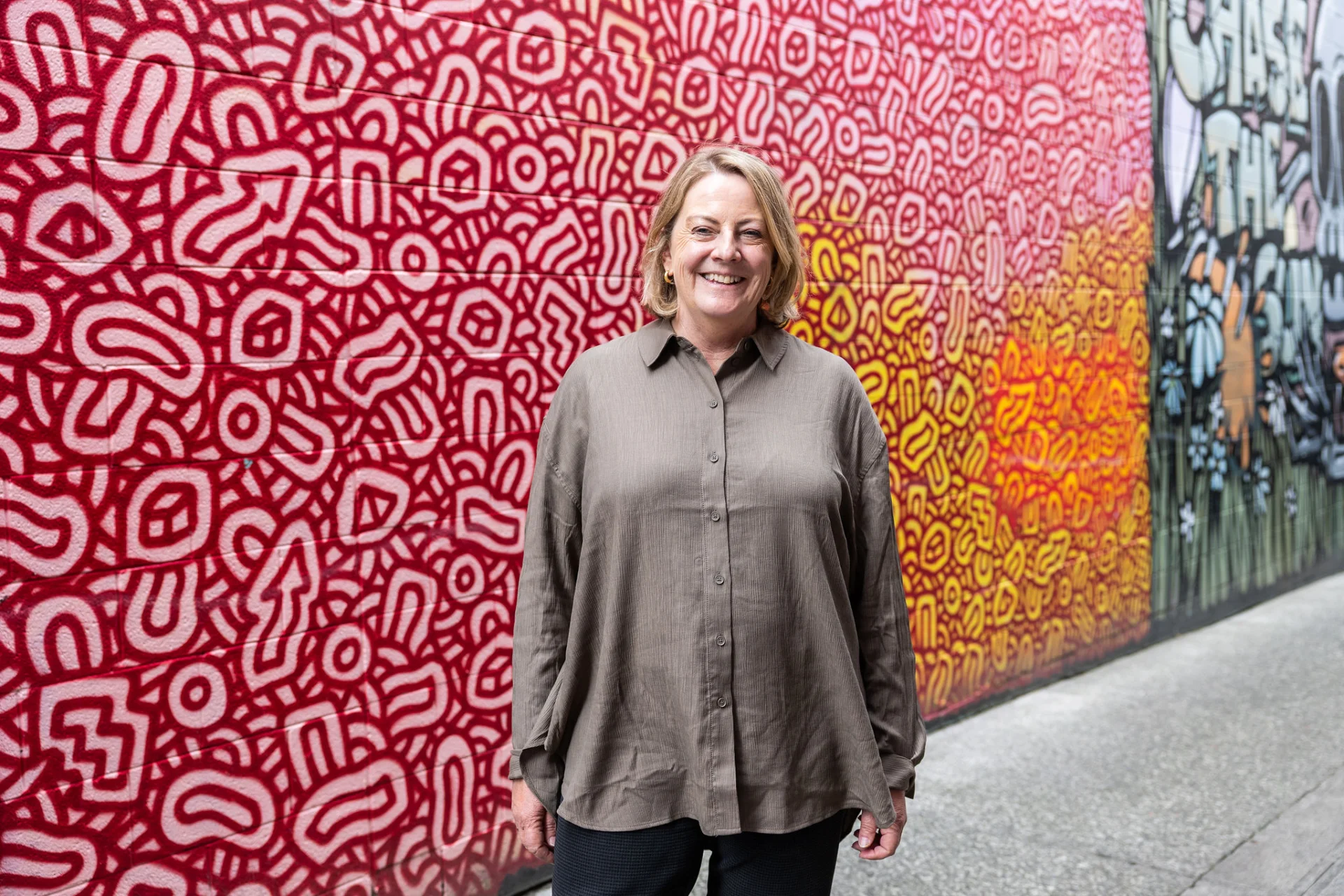 A professional portrait of Janis Levieux standing outdoors in front of a red and yellow mural with abstract patterns. She wears a brown button-up shirt, with short blonde hair, smiling directly at the camera with a warm and friendly expression.