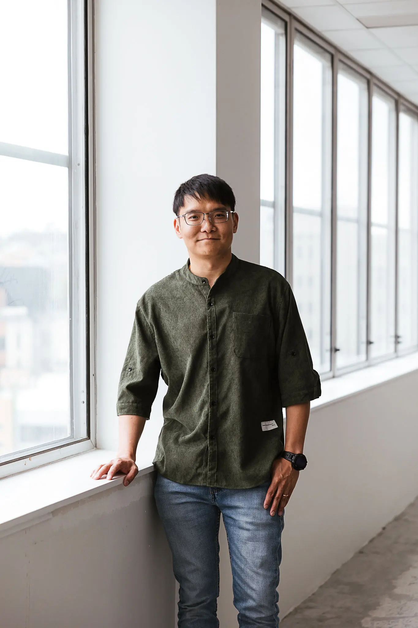 A professional headshot of Jax Cheng standing indoors beside a large window, softly lit by natural light. He wears a dark green button-up shirt with a relaxed collar, with short black hair and glasses, looking directly at the camera with a calm and composed expression.