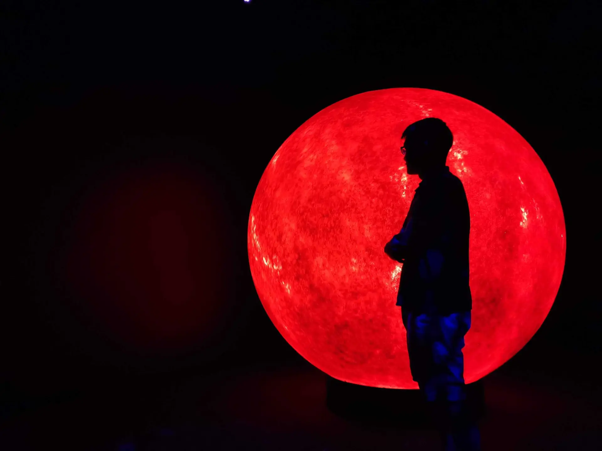 A silhouetted figure standing in front of a large glowing red circular installation in a dark space, the strong backlight defining their outline and posture.