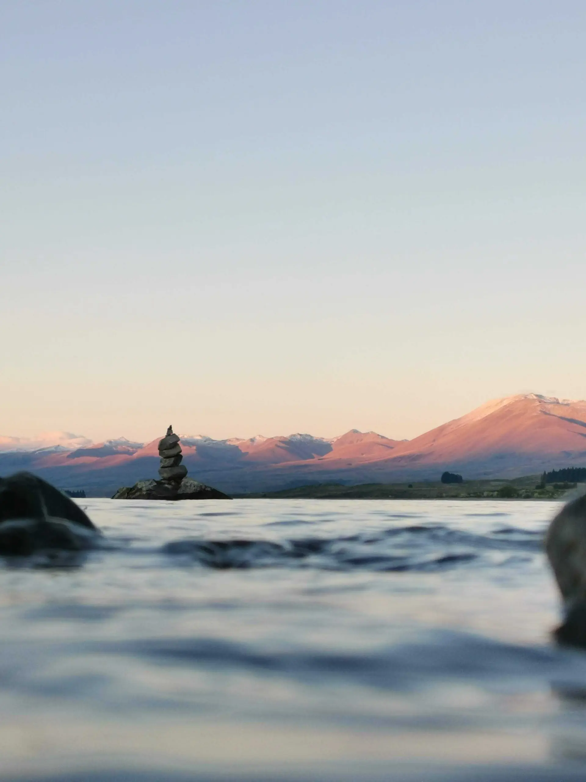A low-angle photograph of a small stack of stones balanced in shallow water, with soft ripples in the foreground and distant mountains catching warm light in the background.