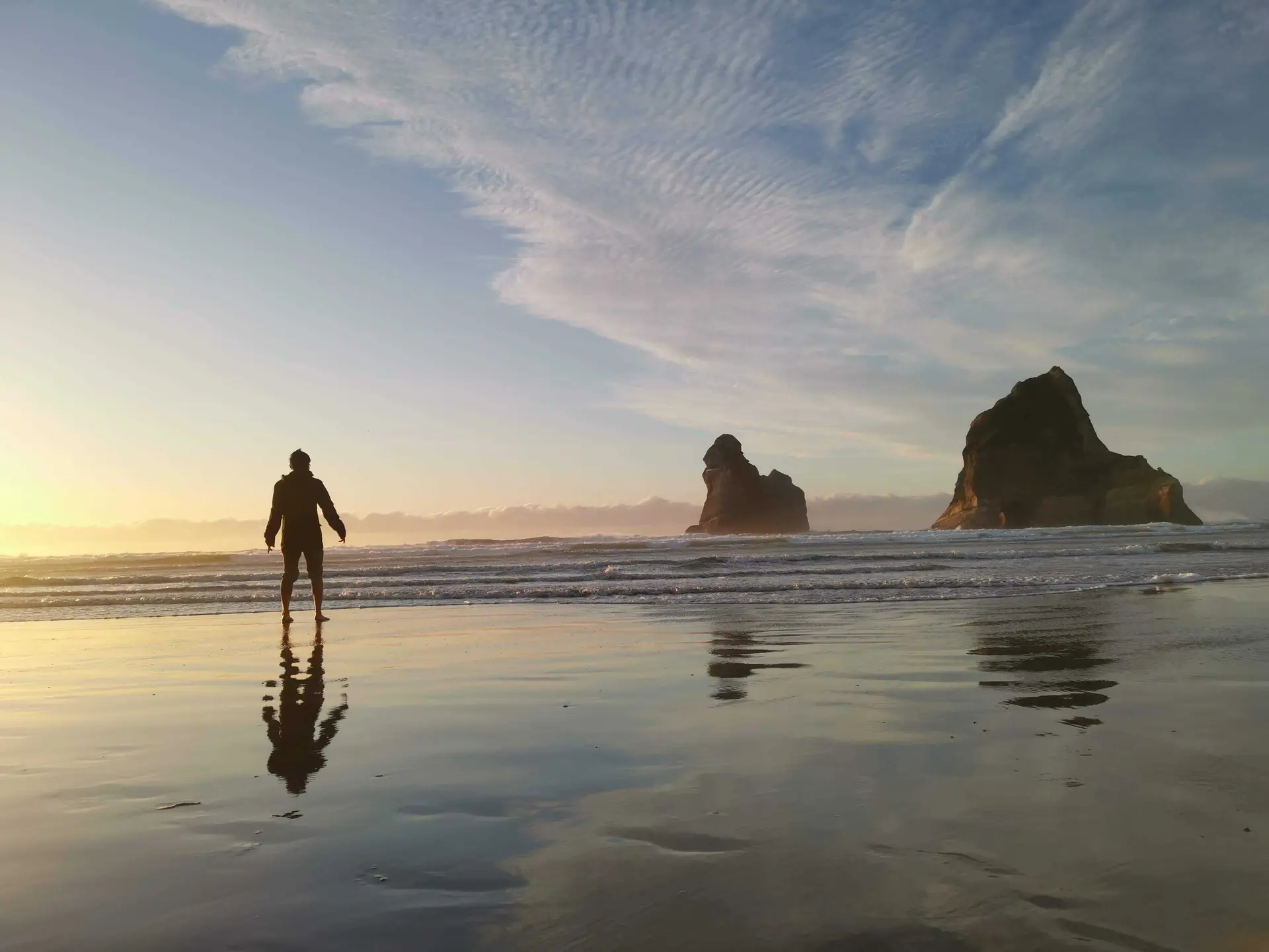 A wide coastal scene showing a person standing on wet sand at sunset, their reflection mirrored in the surface, with large rock formations rising from the ocean in the distance.