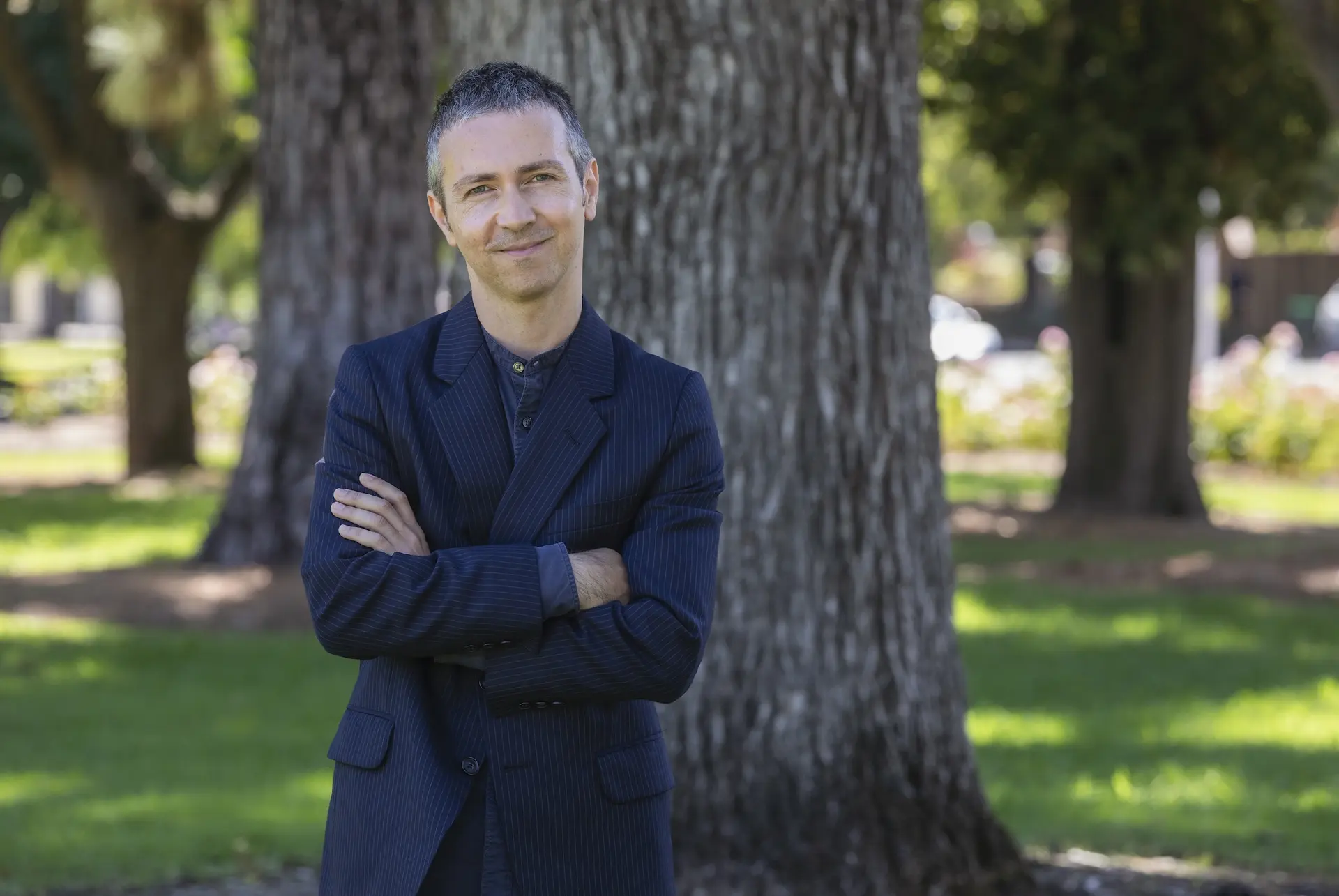 A portrait of Jet O’Rourke smiling and standing outdoors in a park setting, wearing a dark pinstriped suit jacket with a buttoned shirt, arms crossed, with short hair, positioned in front of large tree trunks and greenery.