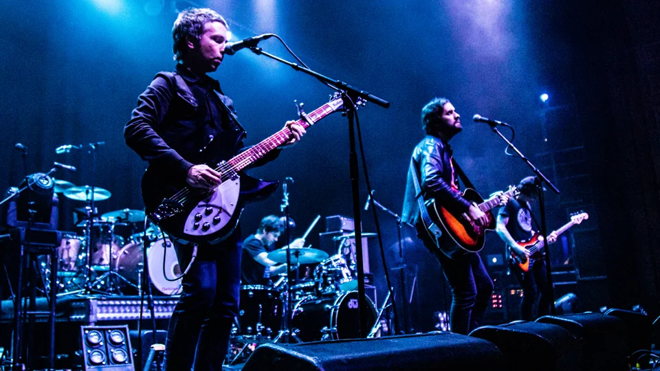 A live performance photograph of Jet playing guitar on stage under blue lighting, accompanied by a band with drums and bass, with microphones and instruments arranged across the stage.