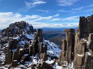 A landscape photograph of a rocky mountain ridge with tall, jagged stone formations dusted with snow, overlooking a wide valley under a bright sky with scattered clouds.