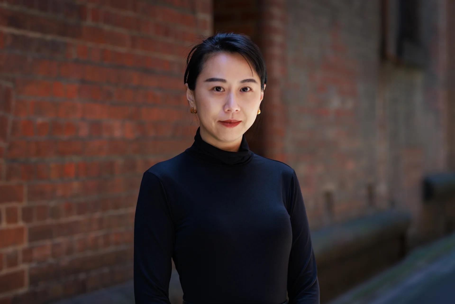 A portrait of Keen Tham standing outdoors against a softly blurred brick wall, wearing a black turtleneck with her hair neatly tied back and small gold earrings, looking directly at the camera smiling with a calm and composed expression.