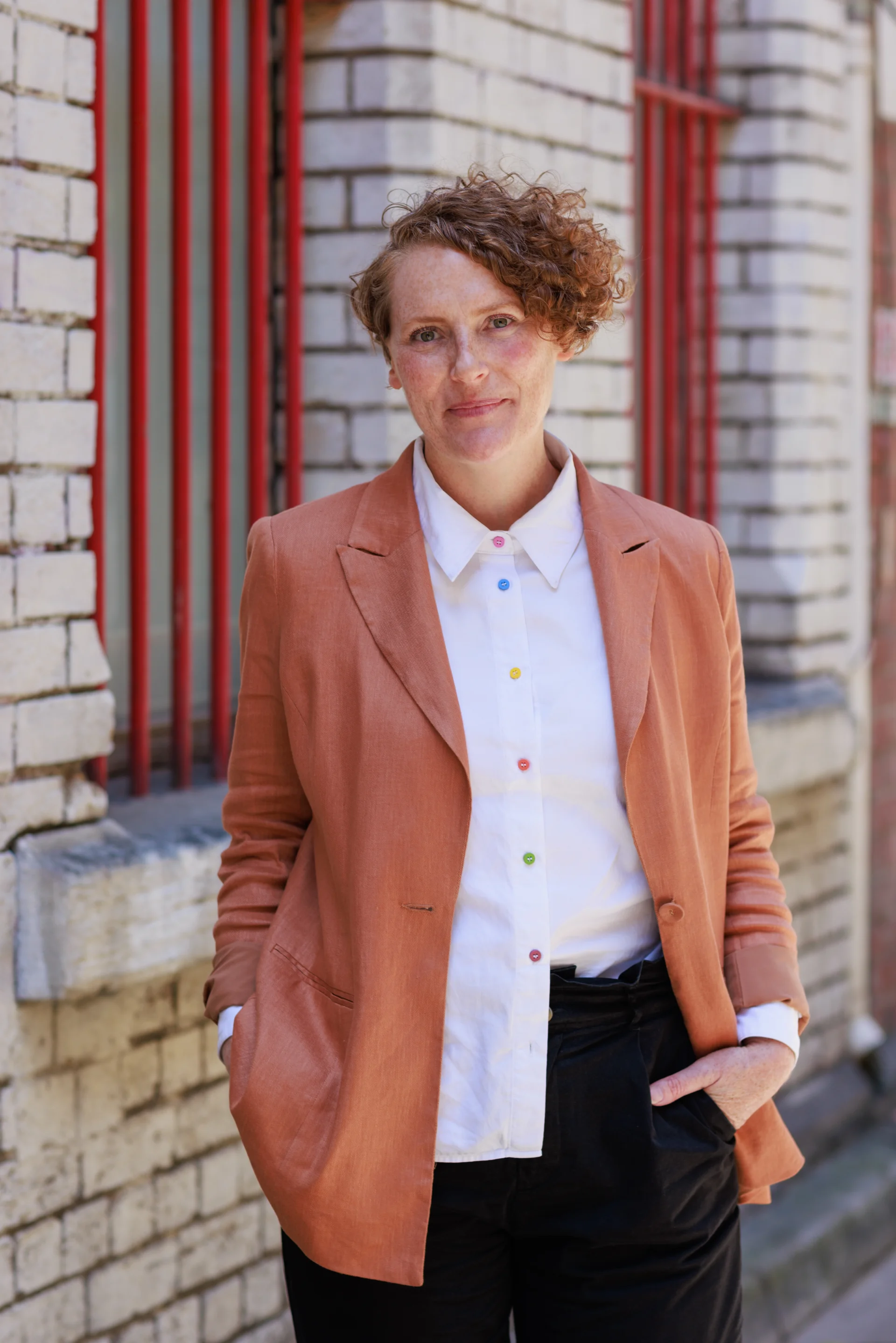 A professional headshot of Kim Sheridan standing against a white brick wall with vertical red window guards. She wears a warm peach-toned blazer over a white shirt with colourful buttons, with short ginger curly hair styled to one side, looking directly at the camera with a composed and approachable expression.
