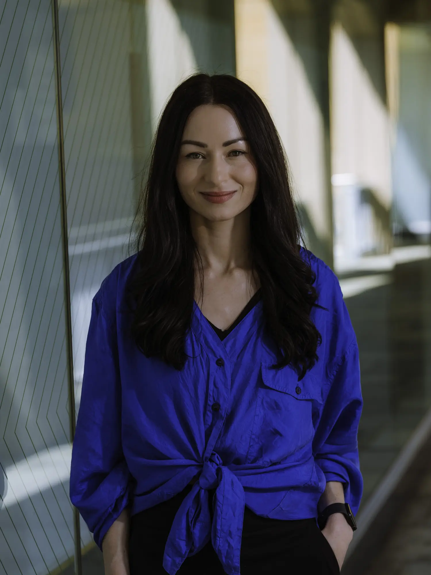 A professional headshot of Kirsten Fox standing indoors beside a glass partition, softly lit by natural light. She wears a blue button-up shirt, with long dark hair worn down, smiling directly at the camera with a warm and composed expression.