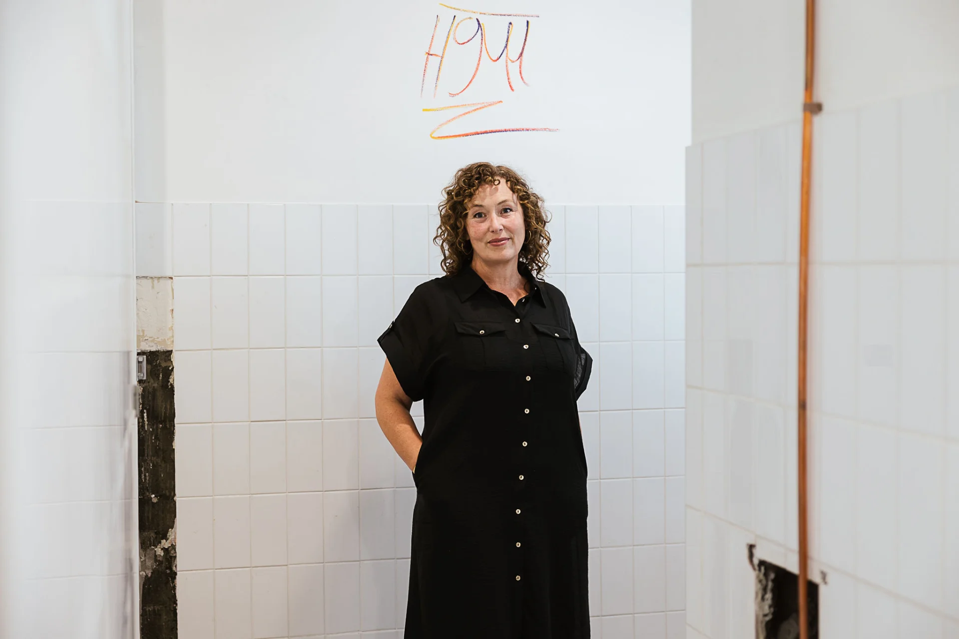 A professional headshot of Kylee Scott standing against a white tiled interior wall with subtle industrial details. She wears a black button-up dress, hands in pockets, looking directly at the camera with a warm and composed expression.