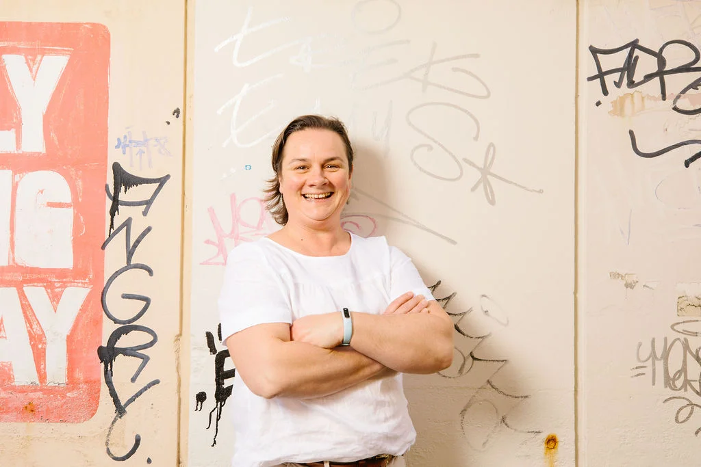 A professional portrait of Kylie Harvey standing outdoors against a graffiti-covered wall, arms crossed and smiling directly at the camera. She wears a light short-sleeve top, with her hair pulled back, lit by soft natural daylight.