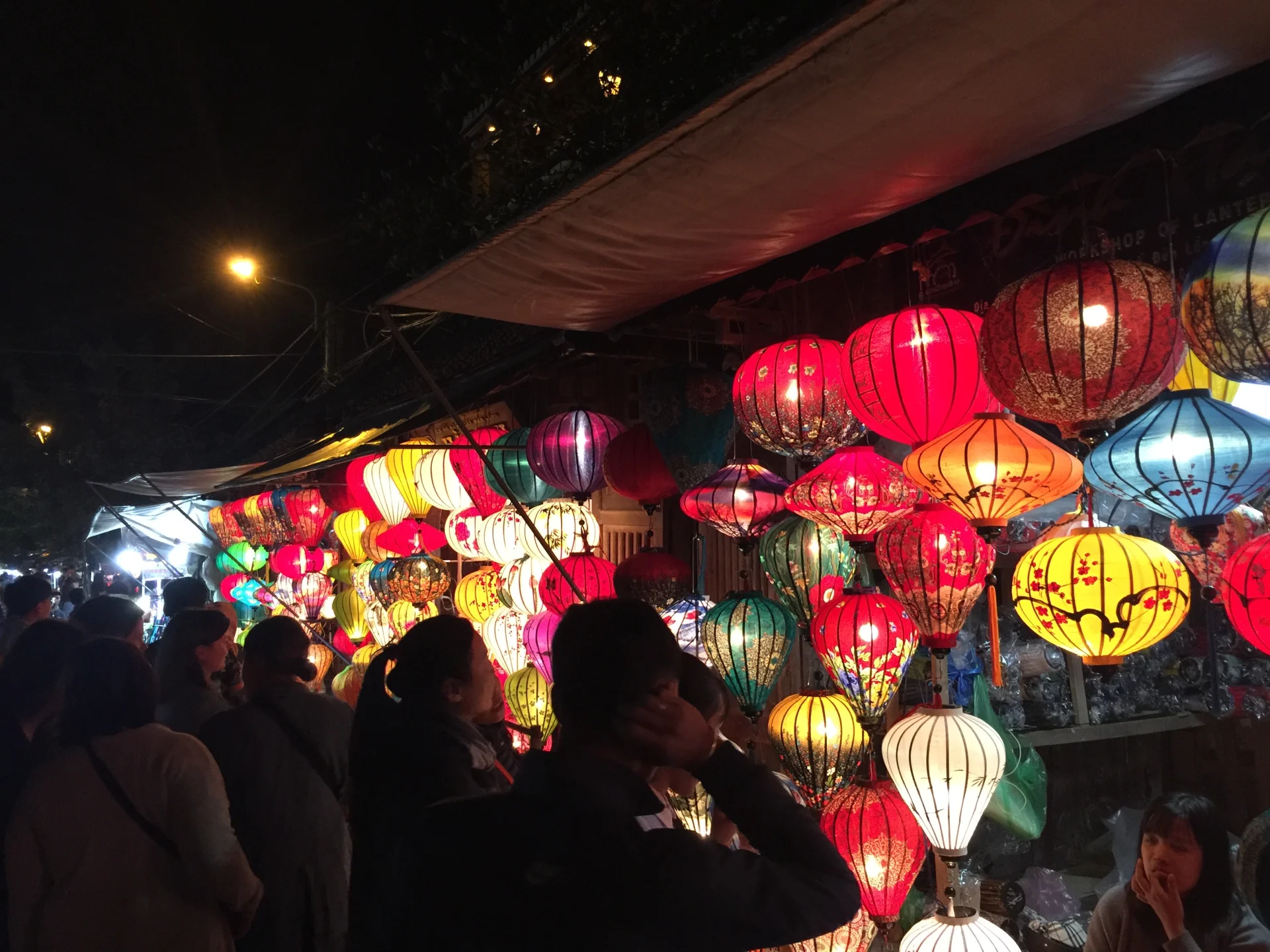 A lively night market scene filled with rows of brightly coloured hanging lanterns in red, yellow, blue, and green, illuminating a crowded street of people browsing stalls beneath a dark sky.
