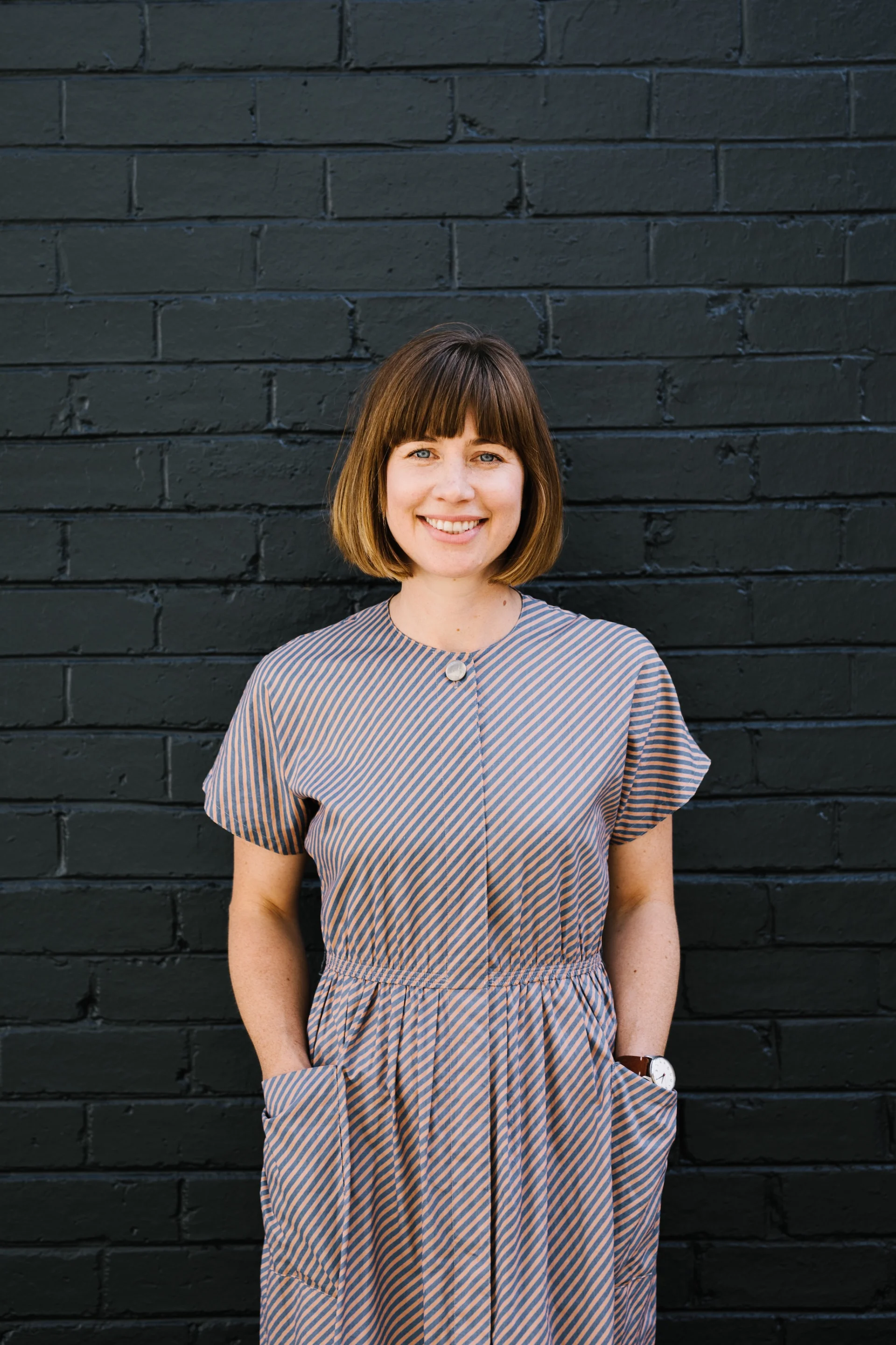 A portrait of Lucia Wellington standing against a dark painted brick wall, smiling directly at the camera. She has a short bob haircut with straight bangs and wears a striped short-sleeve dress.