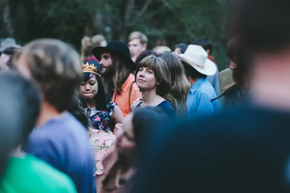 A candid photograph of Lucia Wellington within a crowd at an outdoor gathering, her face in soft focus as she looks slightly upward, surrounded by people in hats and casual clothing.