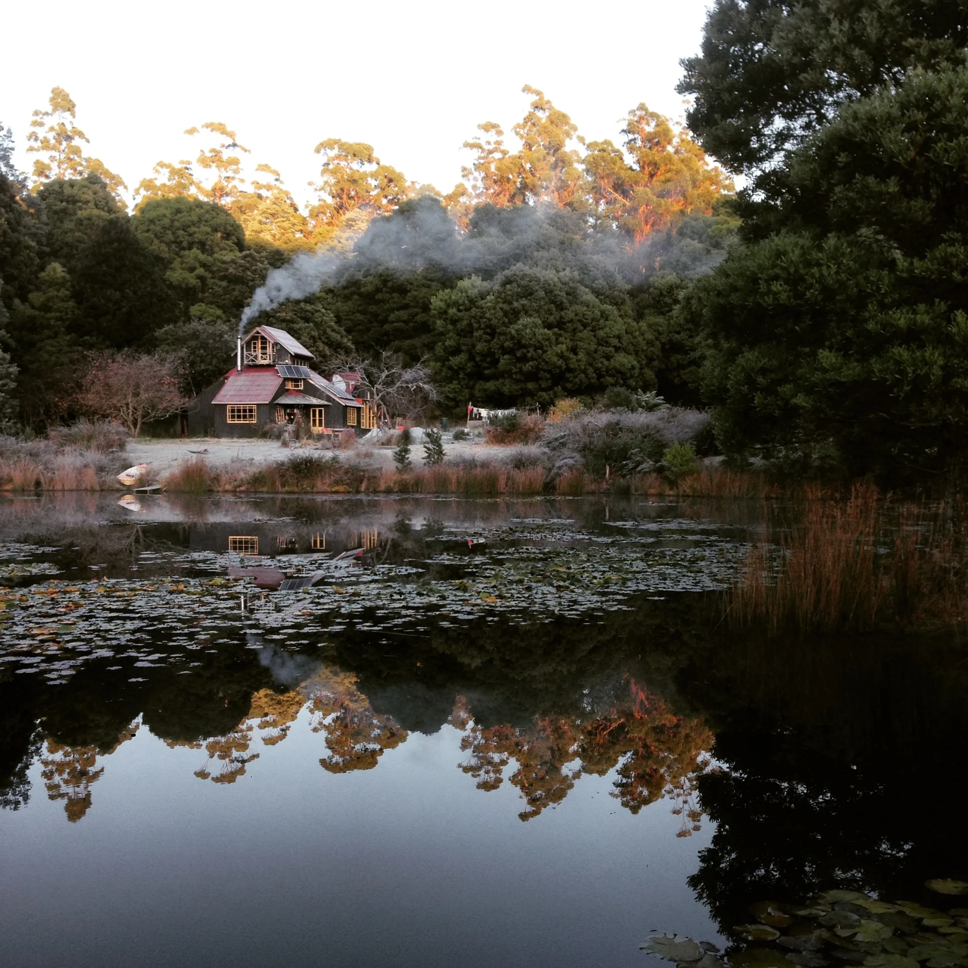 A quiet rural scene of a small timber house nestled among dense trees beside a still pond, with soft smoke rising from the chimney and the building reflected clearly in the water’s surface.