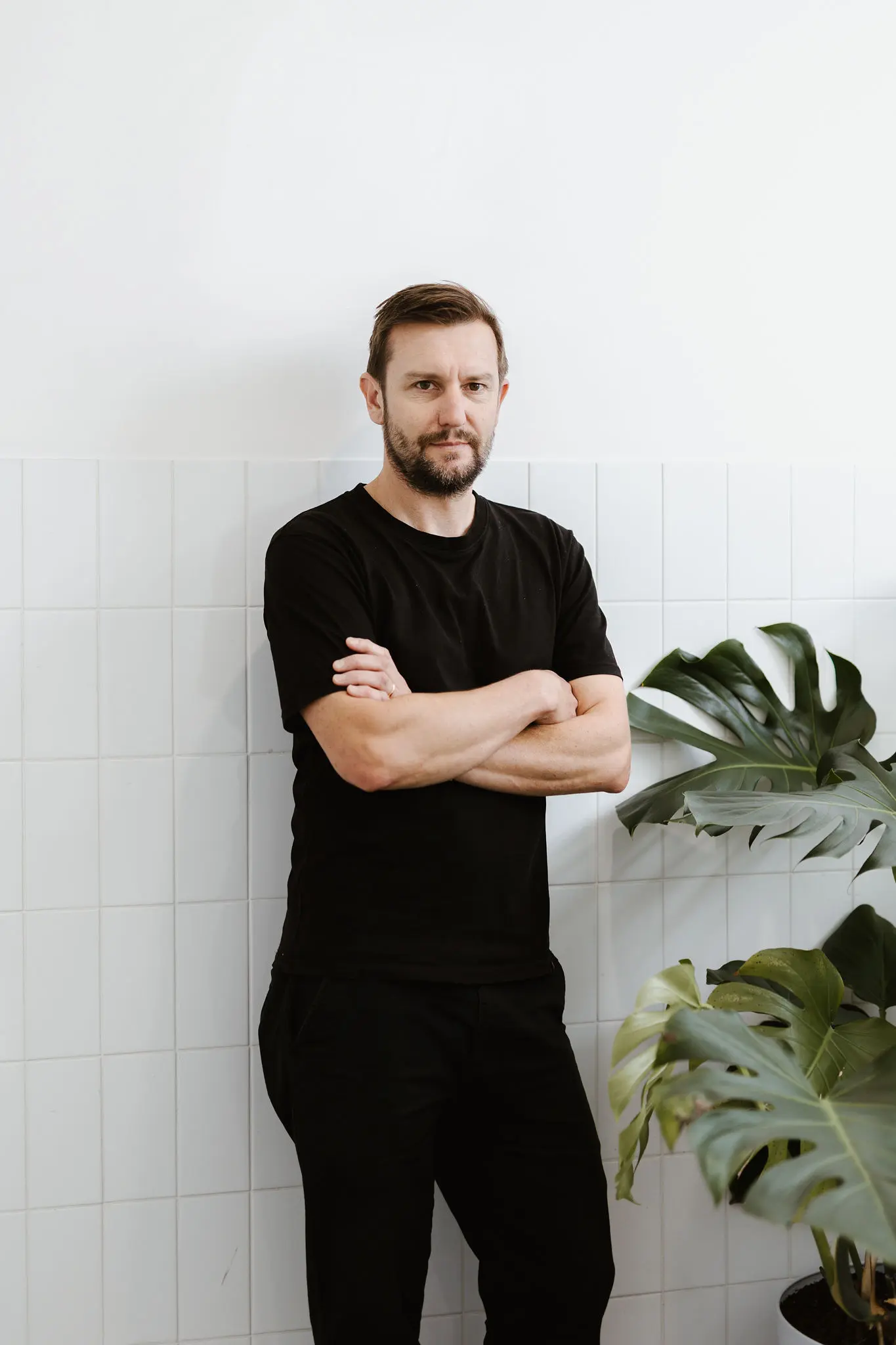 A professional portrait of Luke Waldron standing indoors against a white tiled wall, arms crossed and looking directly at the camera with a friendly, calm expression. He wears a black t-shirt, with short brown hair and beard, softly lit by natural light, with a Monstera plant partially visible to one side.