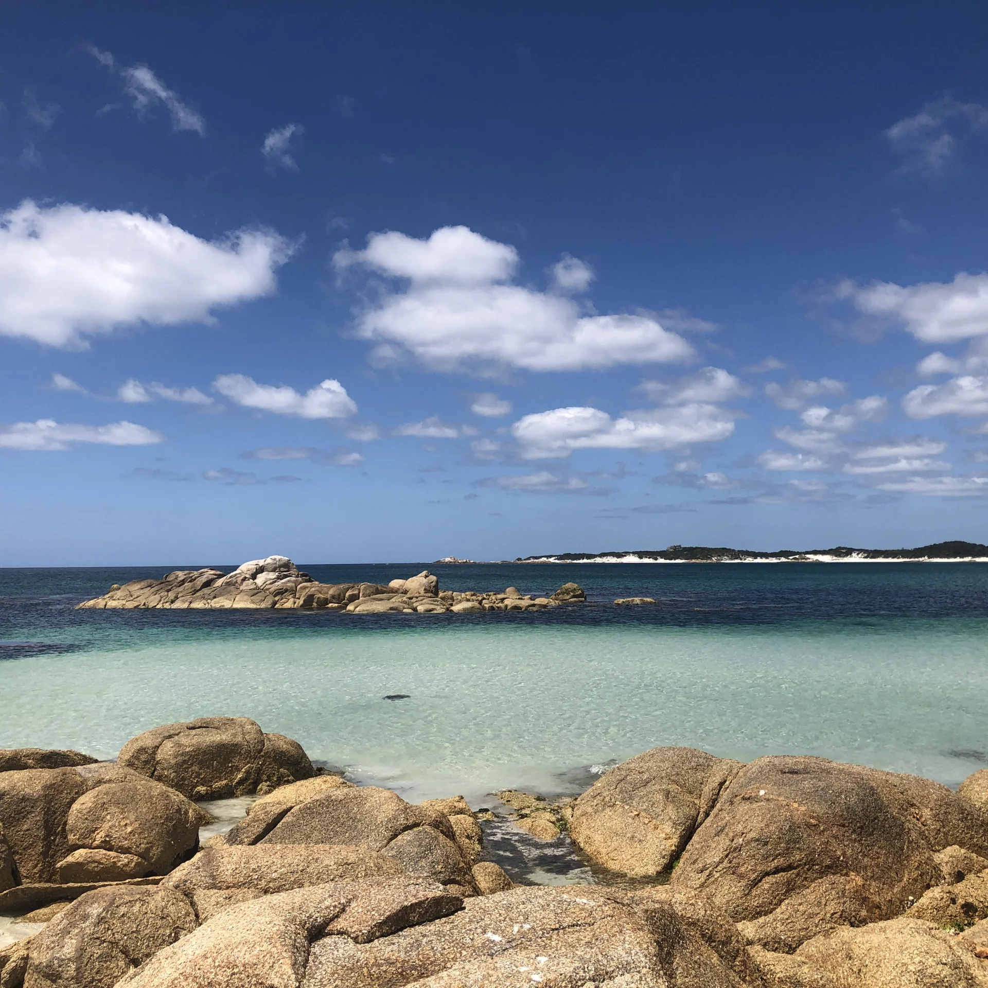 A coastal scene of clear turquoise water meeting a rocky shoreline, with rounded granite boulders in the foreground and a low, sandy coastline stretching across the horizon beneath a bright blue sky with scattered clouds.