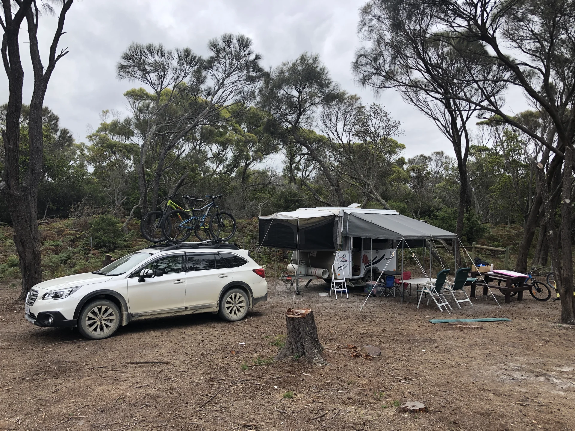 A campsite set within a sparse forest, featuring a parked car with bicycles mounted on top and a camper trailer extended with an awning, surrounded by scattered camping furniture and gear on a dry, earthy ground.
