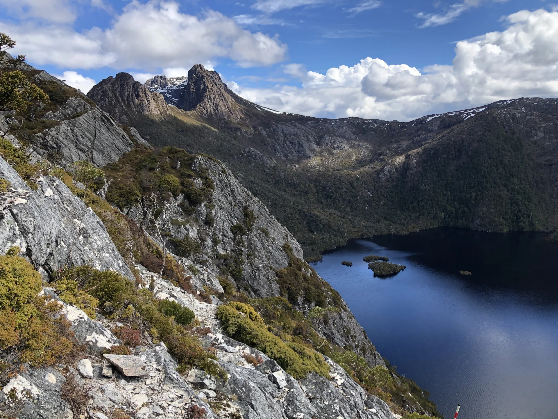 The mountainous landscape at Cradle Mountain, with rugged peaks rising in the distance above Dove Lake below, viewed from a rocky walking track, with patches of vegetation and dramatic cloud formations casting light and shadow across the terrain.
