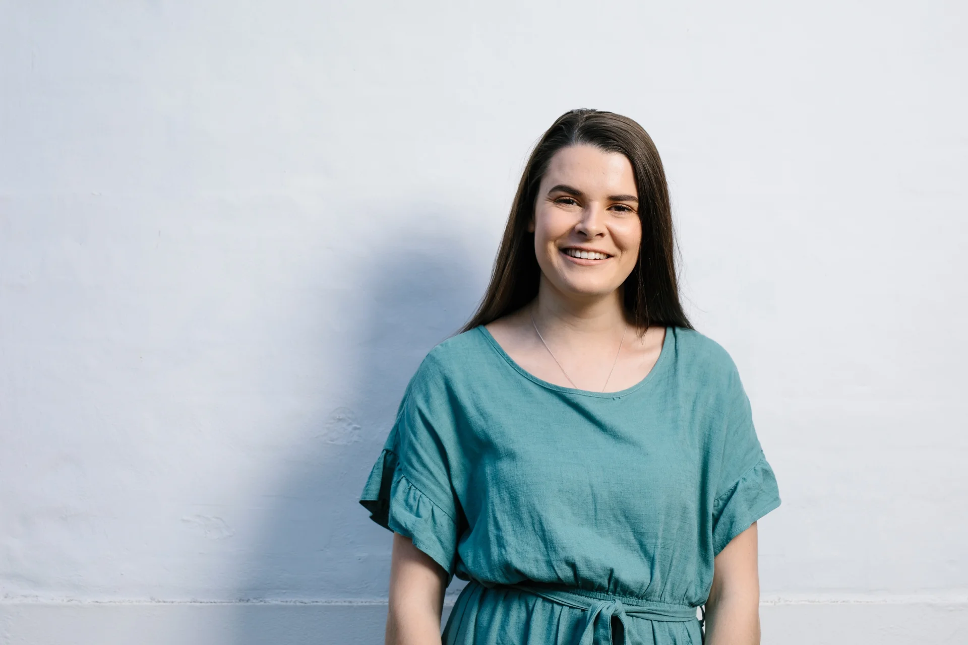 A portrait of Maddie Cooper standing against a plain light wall, smiling warmly at the camera. She wears a loose green dress with short ruffled sleeves, her long dark hair down, and soft natural lighting casts gentle shadows across the background.